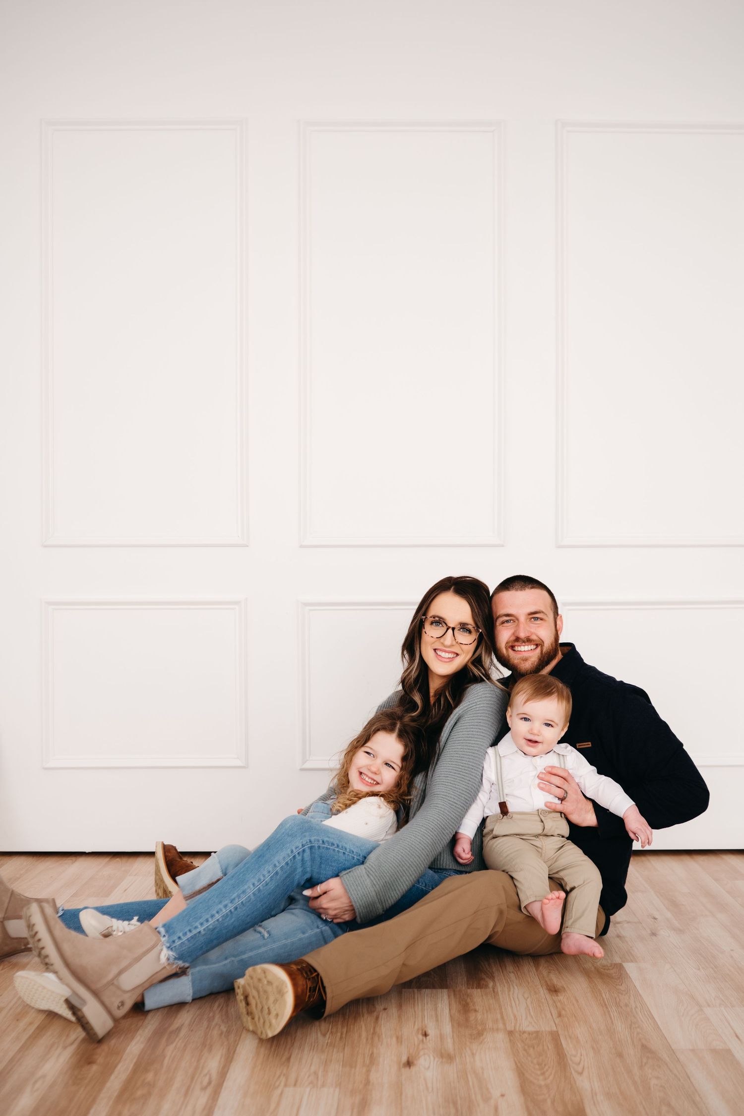 Family sitting together on hardwood floor against white wall wearing casual clothing in studio setting.