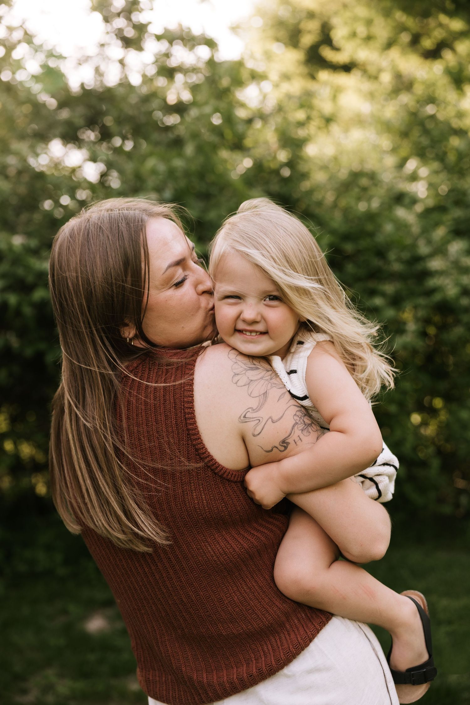 A mother kisses her smiling toddler daughter on the cheek while holding her in a lush green outdoor setting.