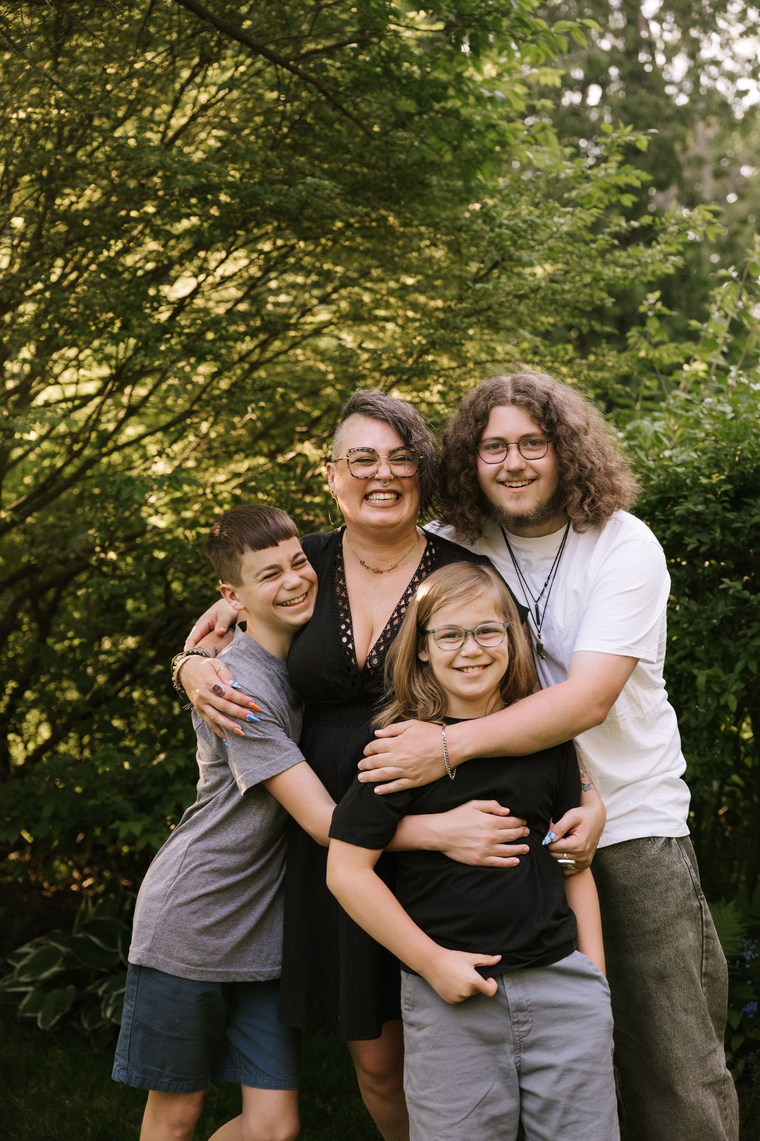 Family of four smiling and hugging outdoors surrounded by lush green trees during golden hour.