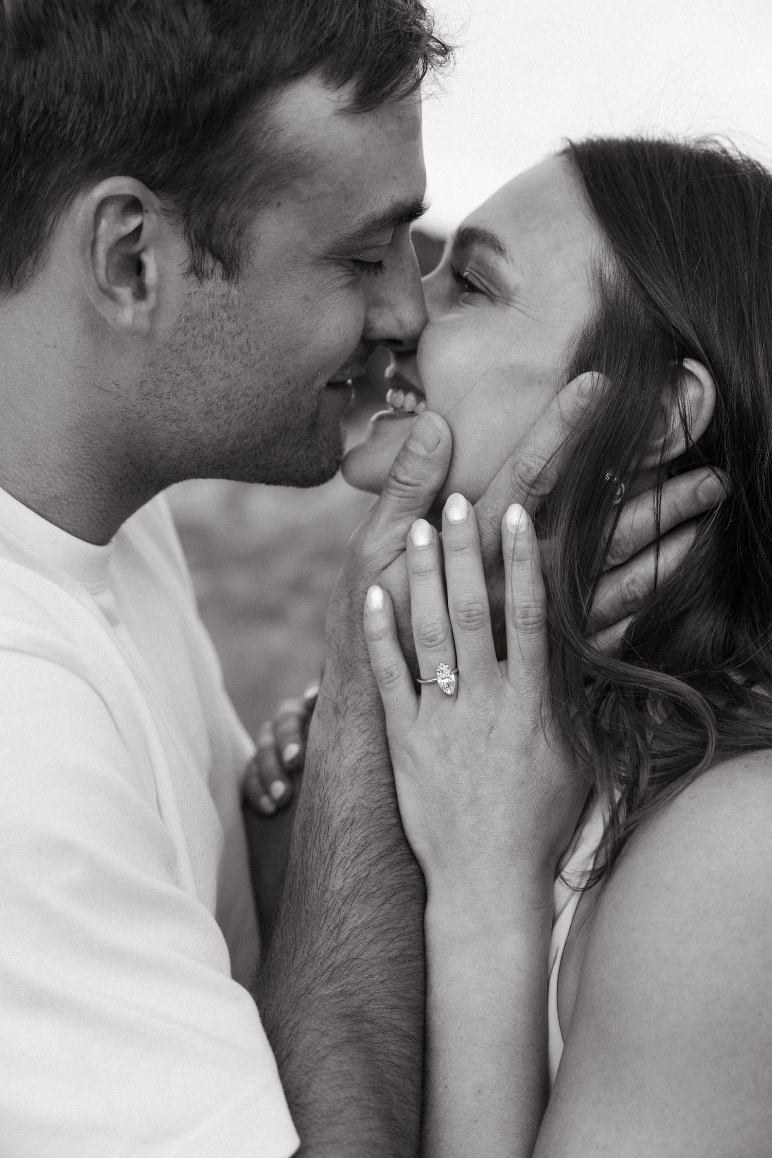 Black and white photo of a smiling couple about to kiss, woman showing off her engagement ring.