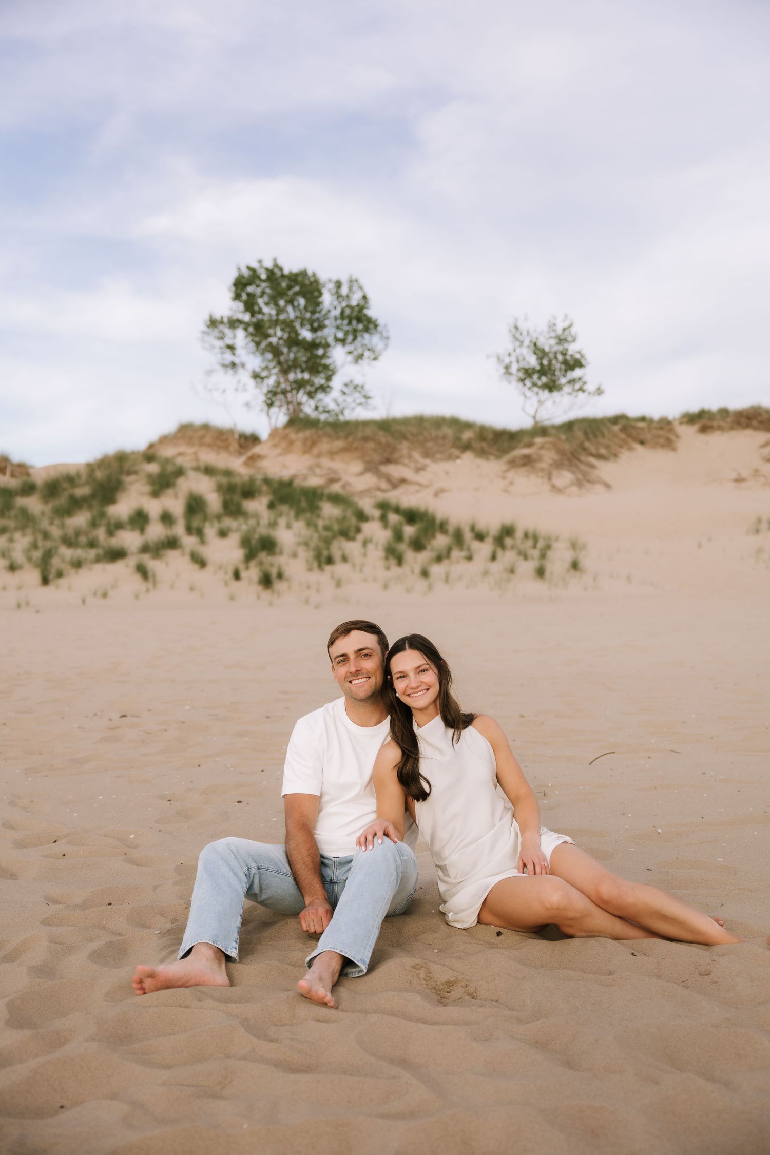 Happy couple sitting together on sandy beach dunes with trees in background under partly cloudy sky.