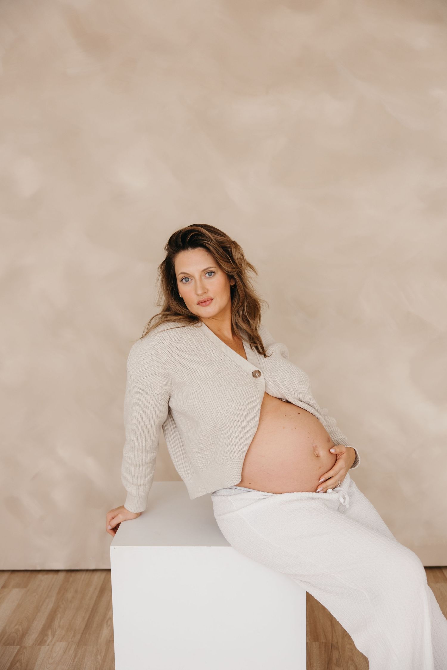 Pregnant woman in a beige knit cardigan sits on a white cube, gently holding her bare baby bump against a beige backdrop.