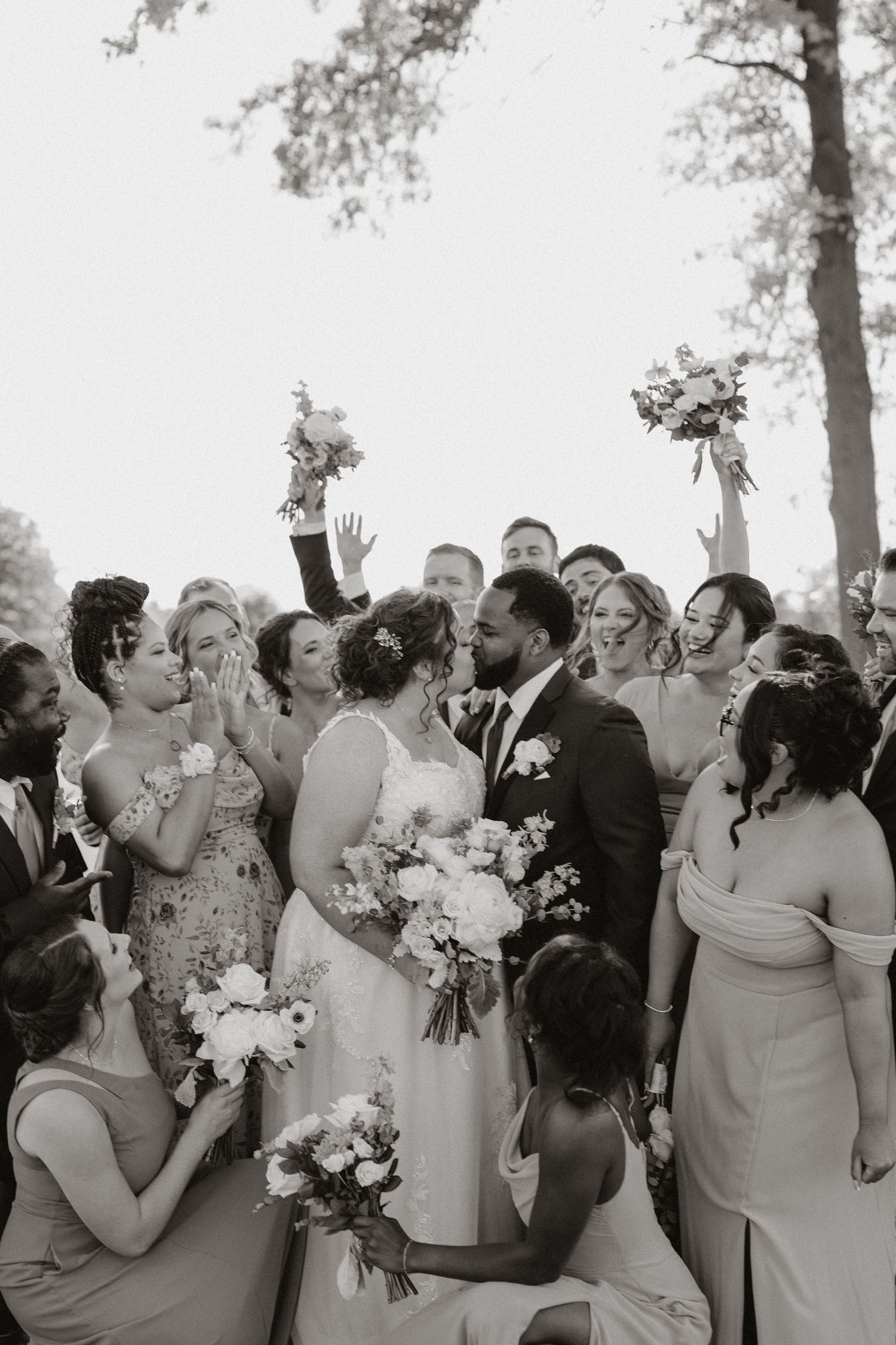 Wedding party celebrating joyfully outdoors with raised bouquets and enthusiastic expressions.