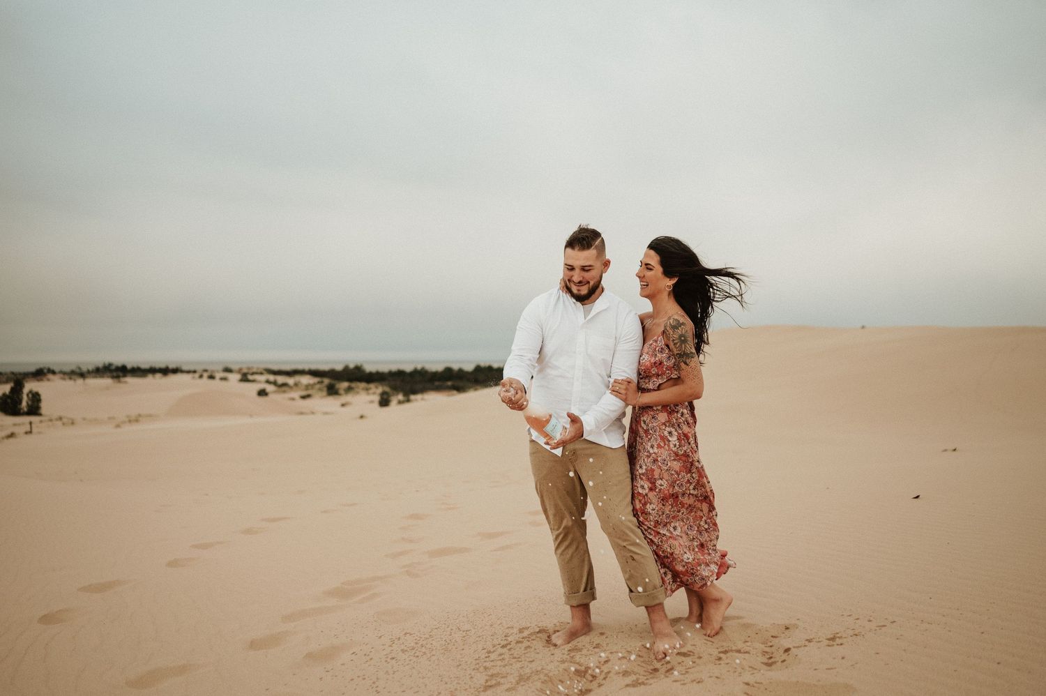 A couple walks hand in hand across windswept sand dunes during a romantic desert photoshoot.