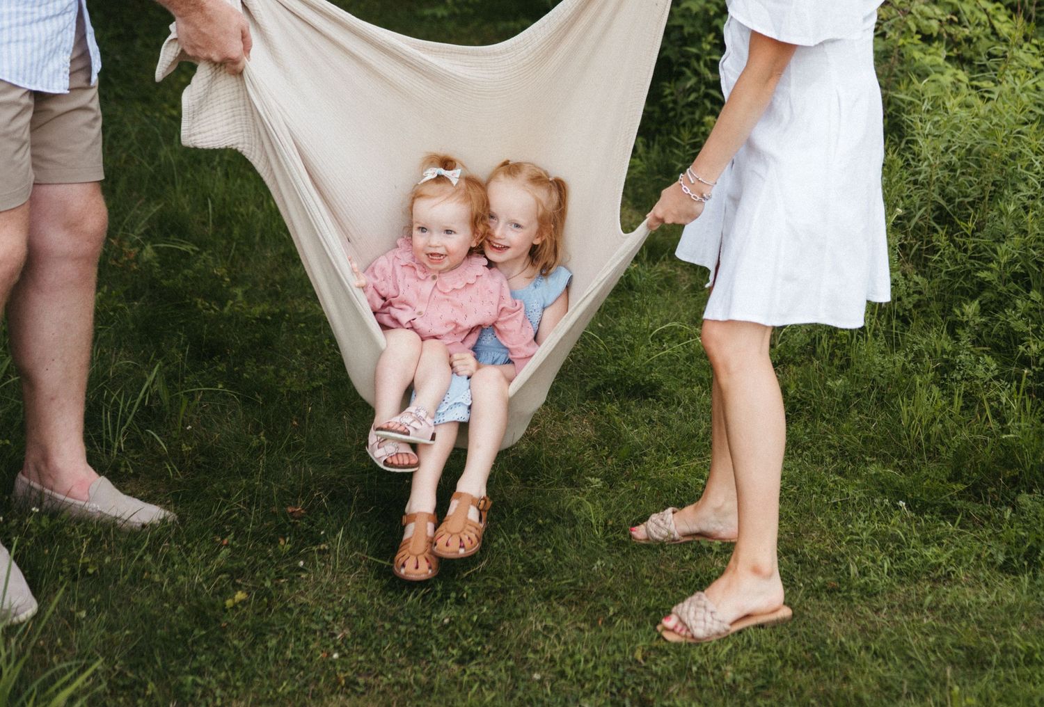 Children swing in a white blanket held by adults in an outdoor garden setting.