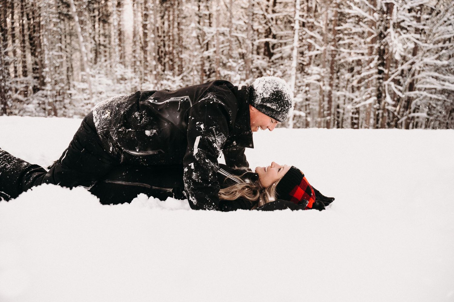 Two people in winter attire laying together in fresh snow surrounded by trees.