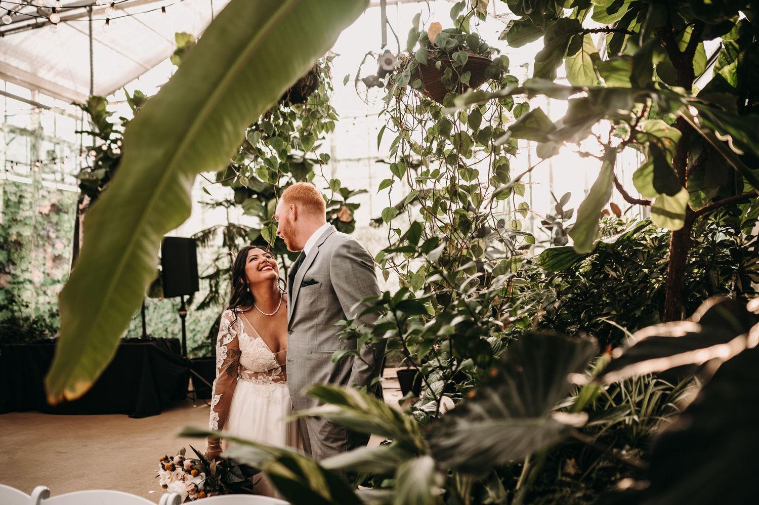 Wedding couple poses in sunlit greenhouse surrounded by lush tropical plants.