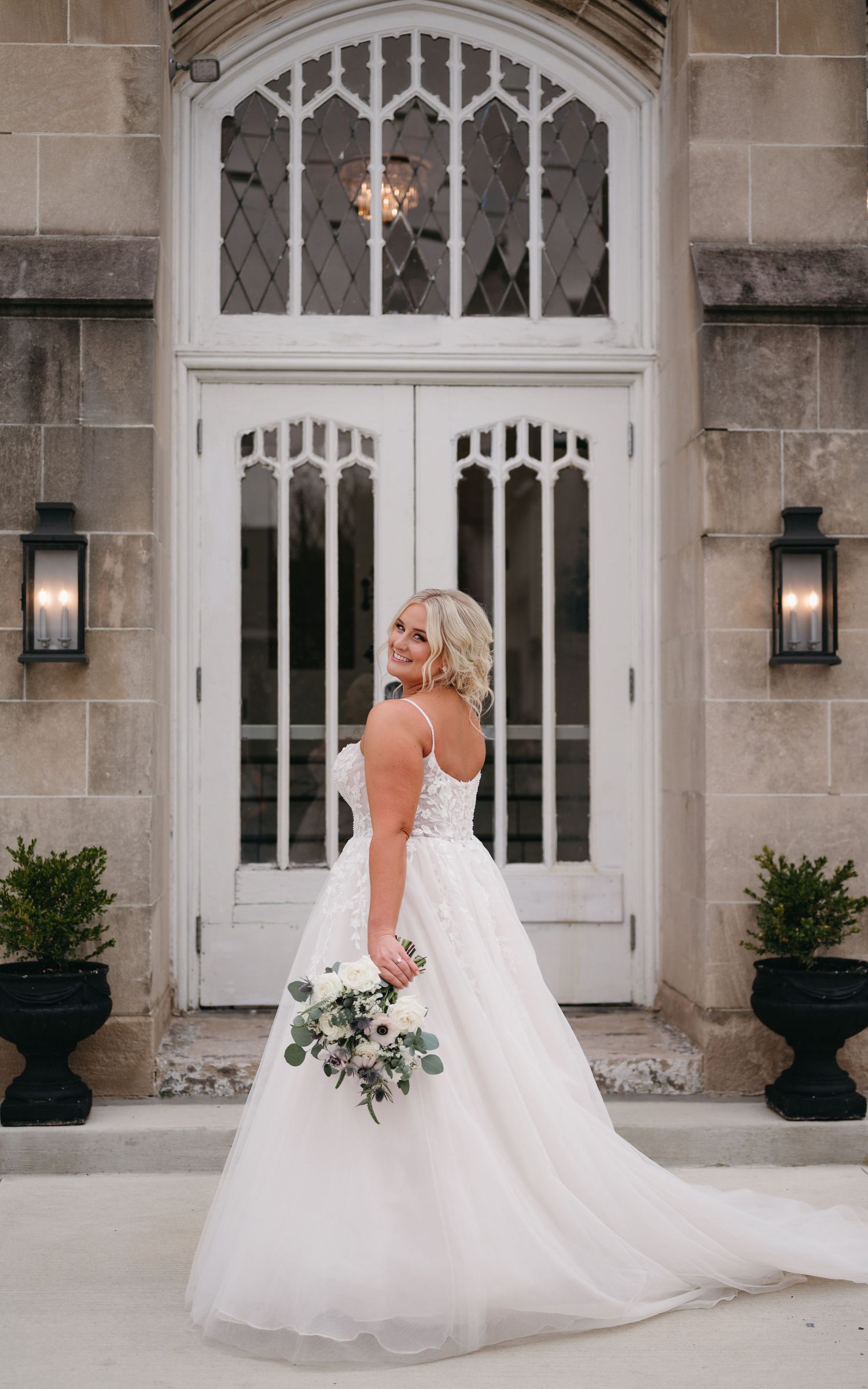 Bride in white wedding gown poses in front of gothic church doorway with bouquet.