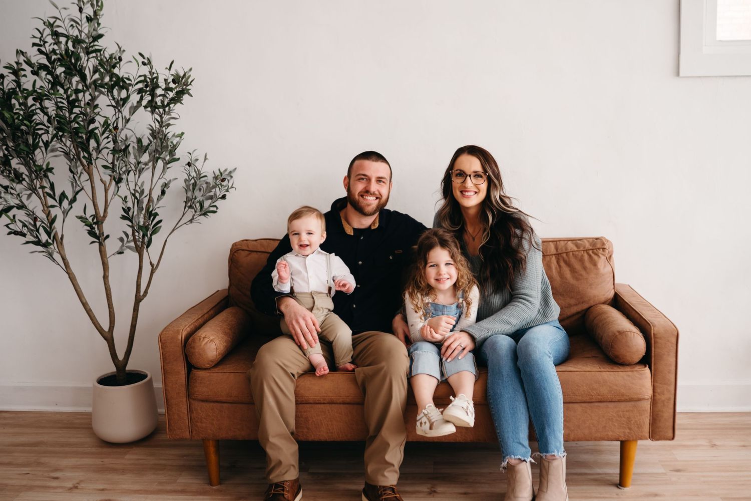 A family of four sits together on a brown leather sofa in a minimal living room with a potted tree.