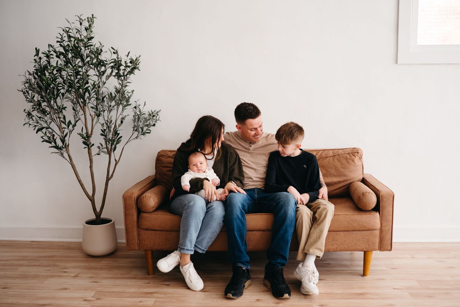 Family sitting together on a brown leather couch in a minimalist living room with a potted olive tree.