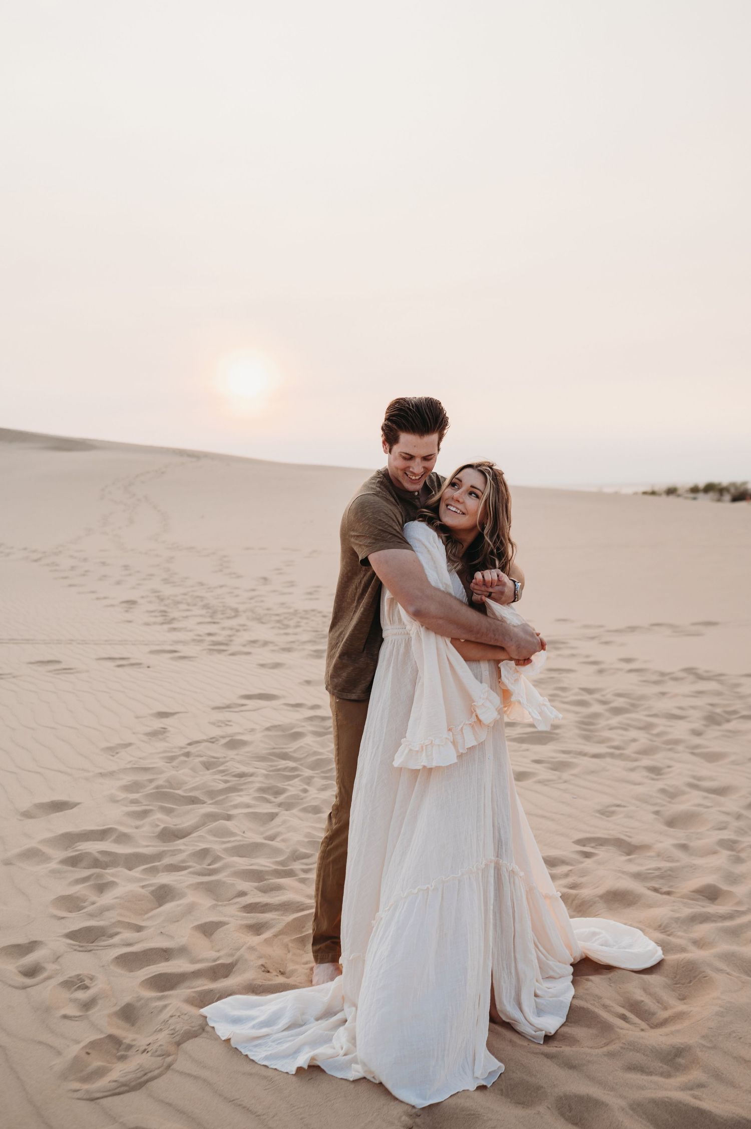 A couple embraces on desert sand during sunset wearing formal attire.