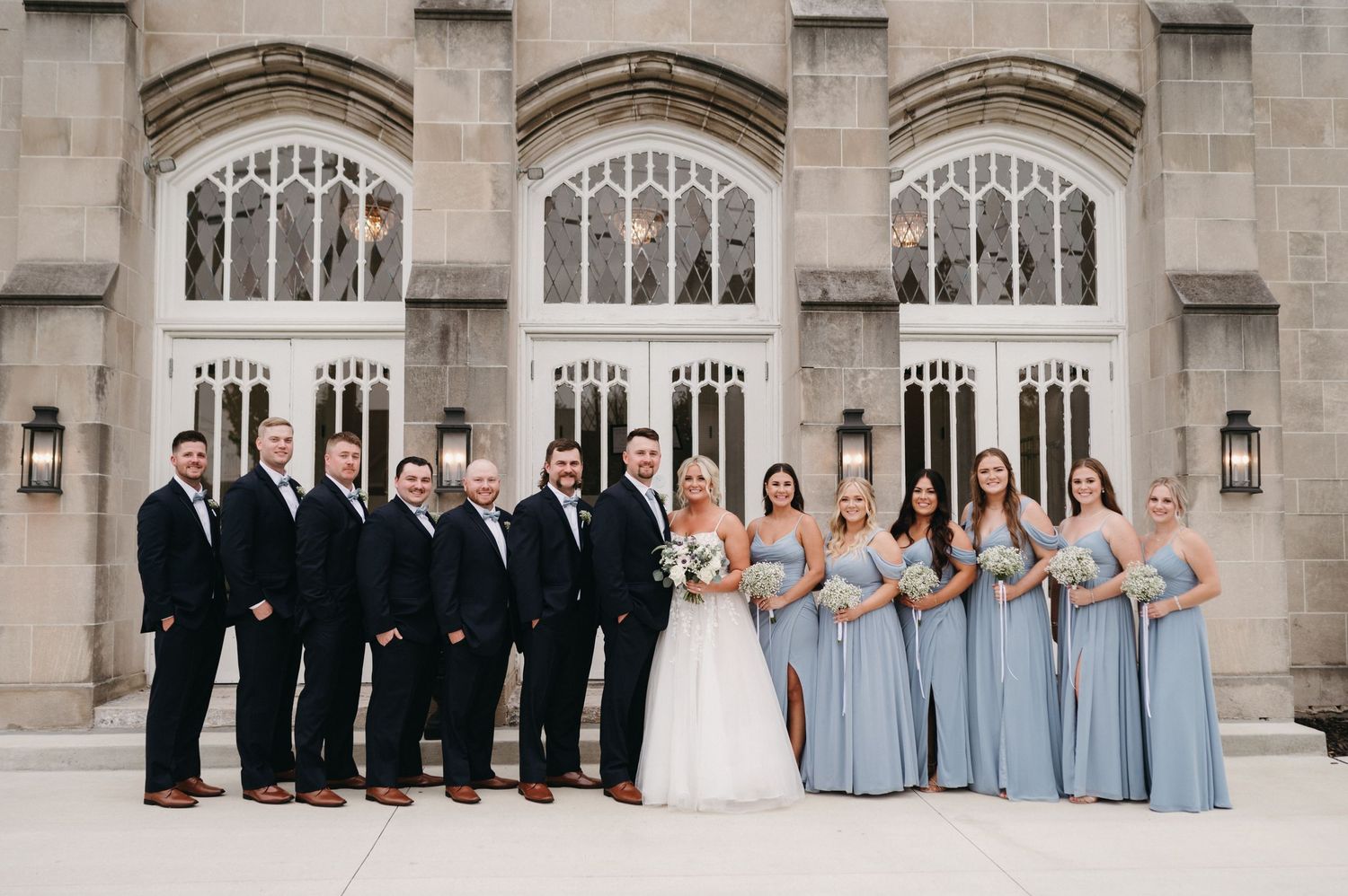 Full wedding party in blue dresses and black tuxedos poses formally against church facade.