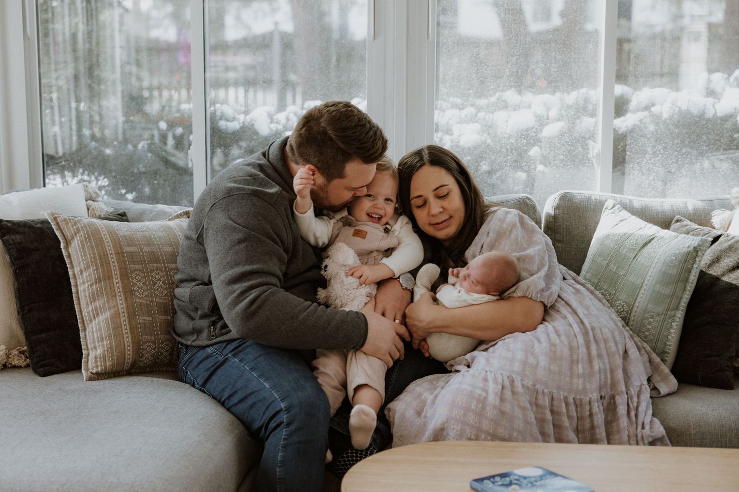 Family cuddles together on grey couch near bright window with sheer curtains.