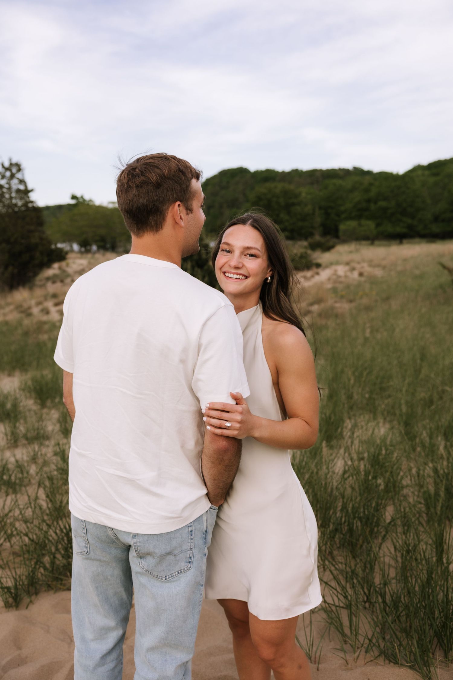 Smiling woman in white mini dress embraces man in white tee amid sandy dunes and green trees.
