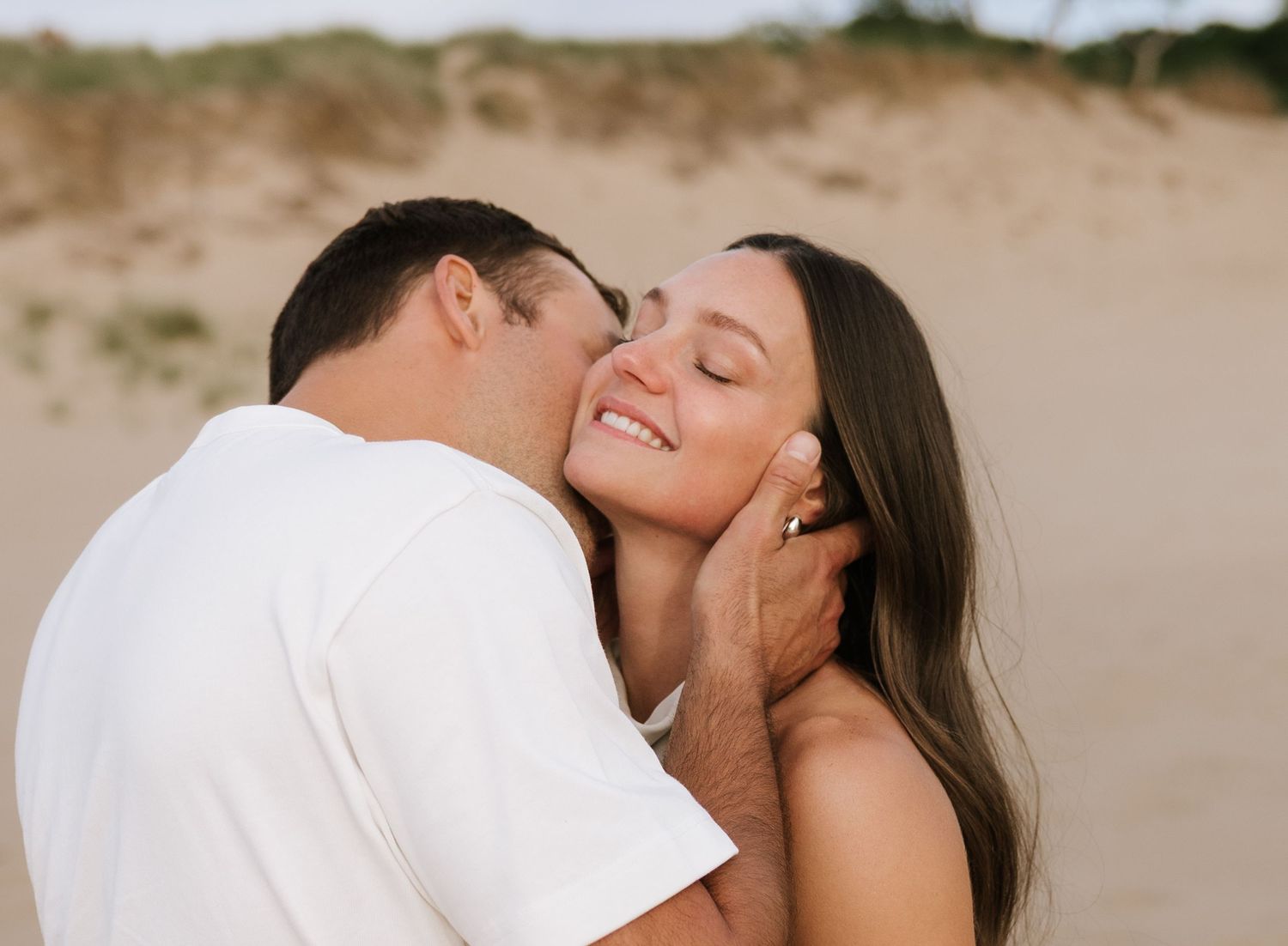 Romantic couple sharing a tender kiss on a sandy beach, woman smiling joyfully in a warm outdoor setting.