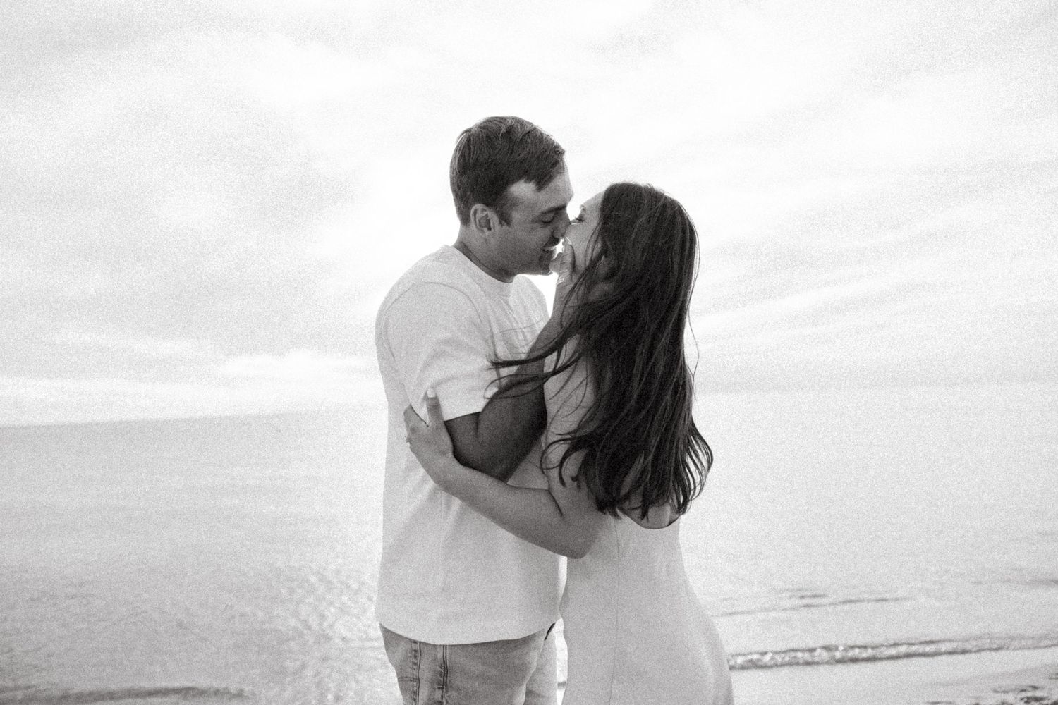 Black and white photo of couple sharing romantic kiss near ocean shoreline, woman's hair blowing in wind.