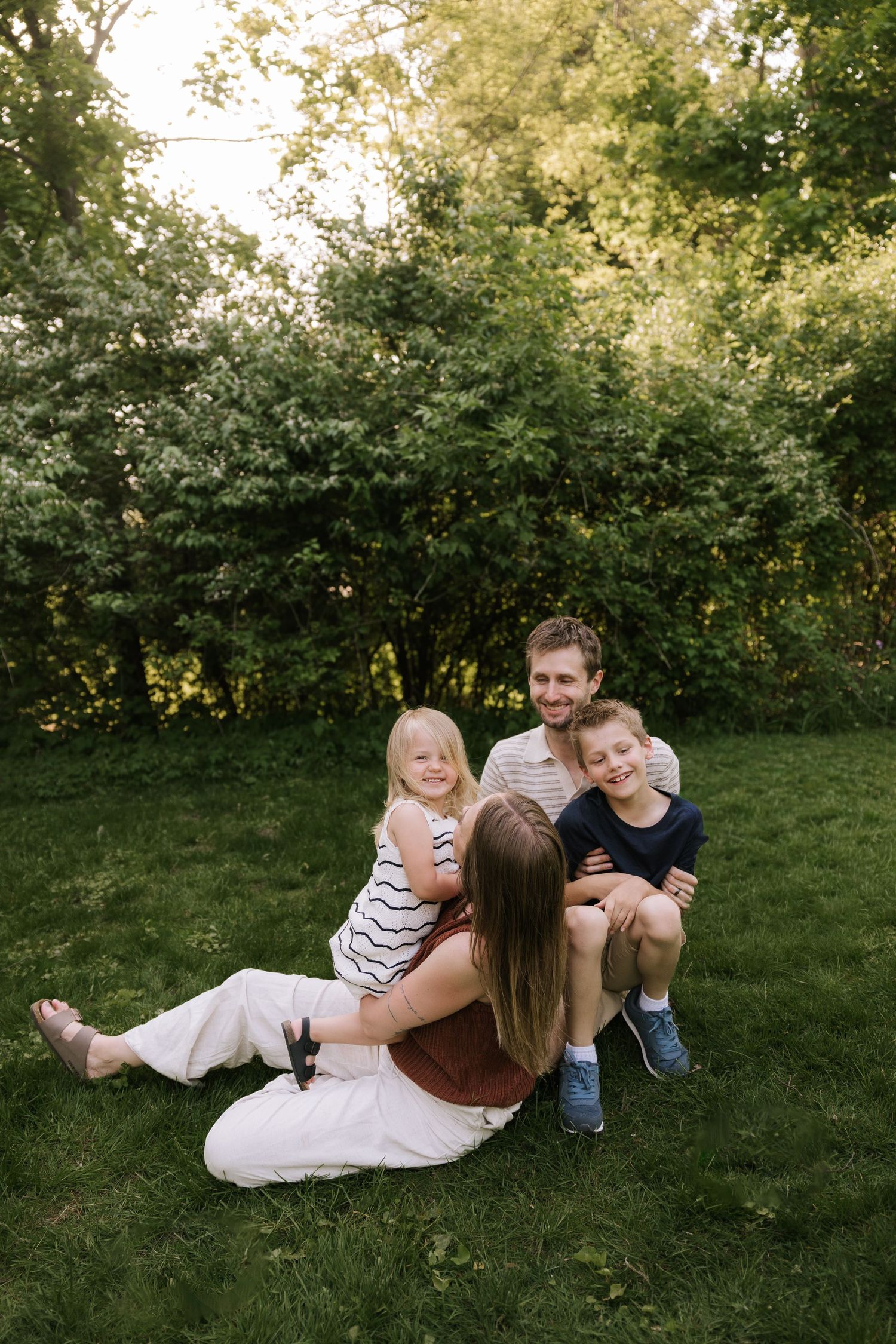 Happy family of four sitting together on green grass outdoors, smiling and enjoying time together near leafy trees.
