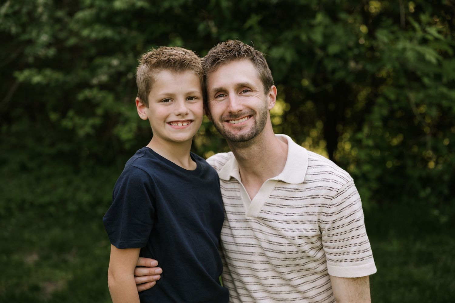 A smiling man and young boy pose together outdoors with lush green trees in the background.