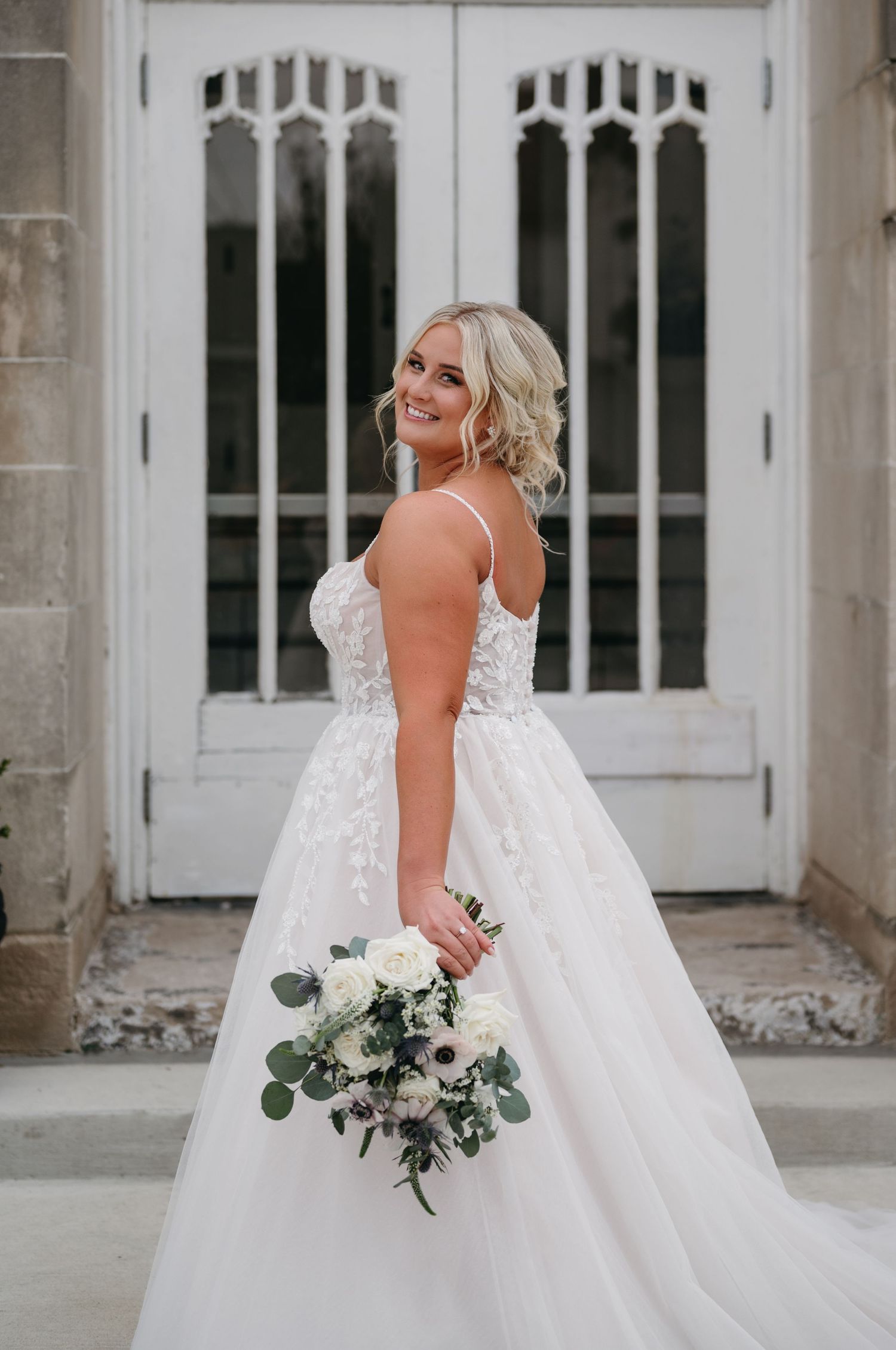 Side view of bride in lace wedding dress holding bouquet against ornate white doors.