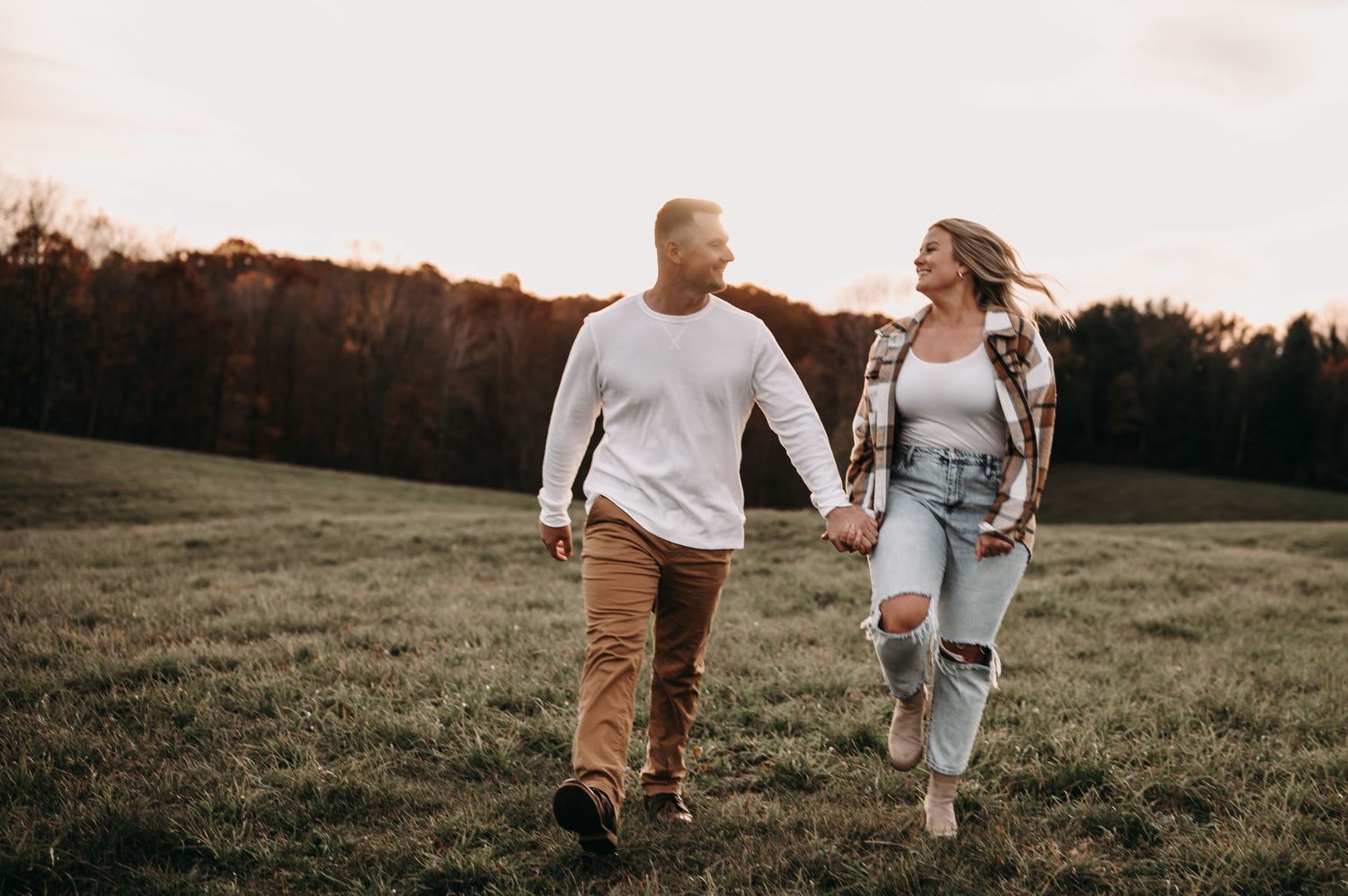Couple laughing while walking together through golden meadow at dusk.