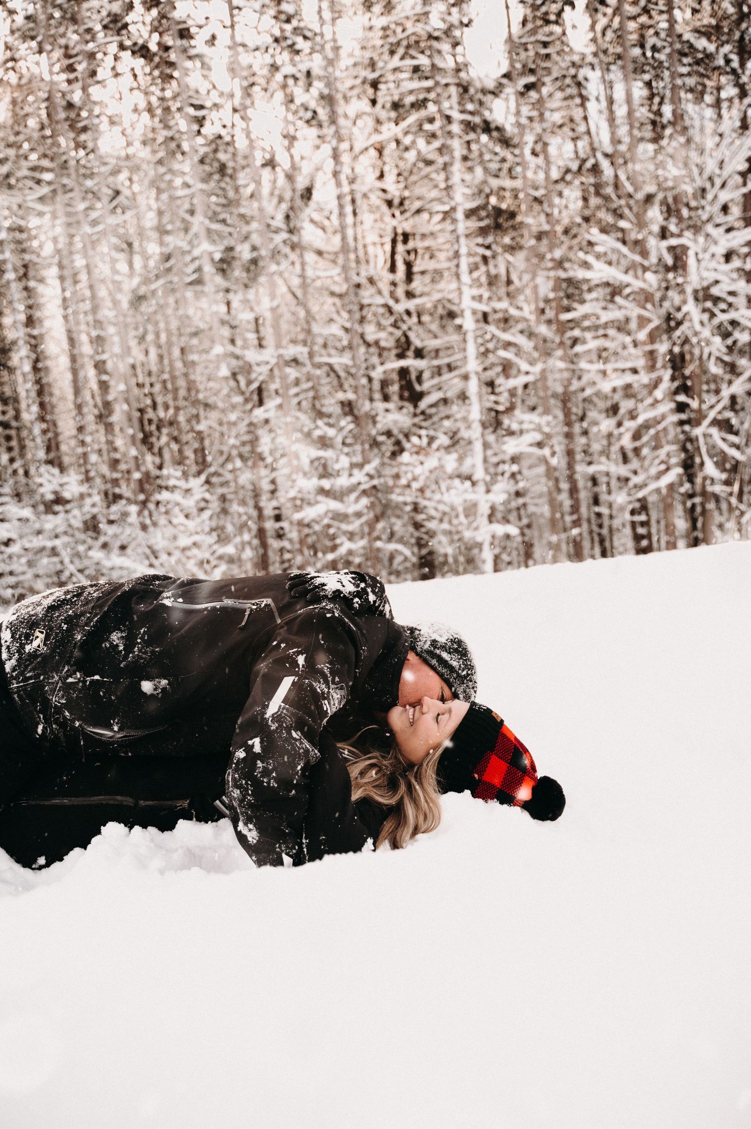 A couple in black winter clothing lying in snow with a forest backdrop.