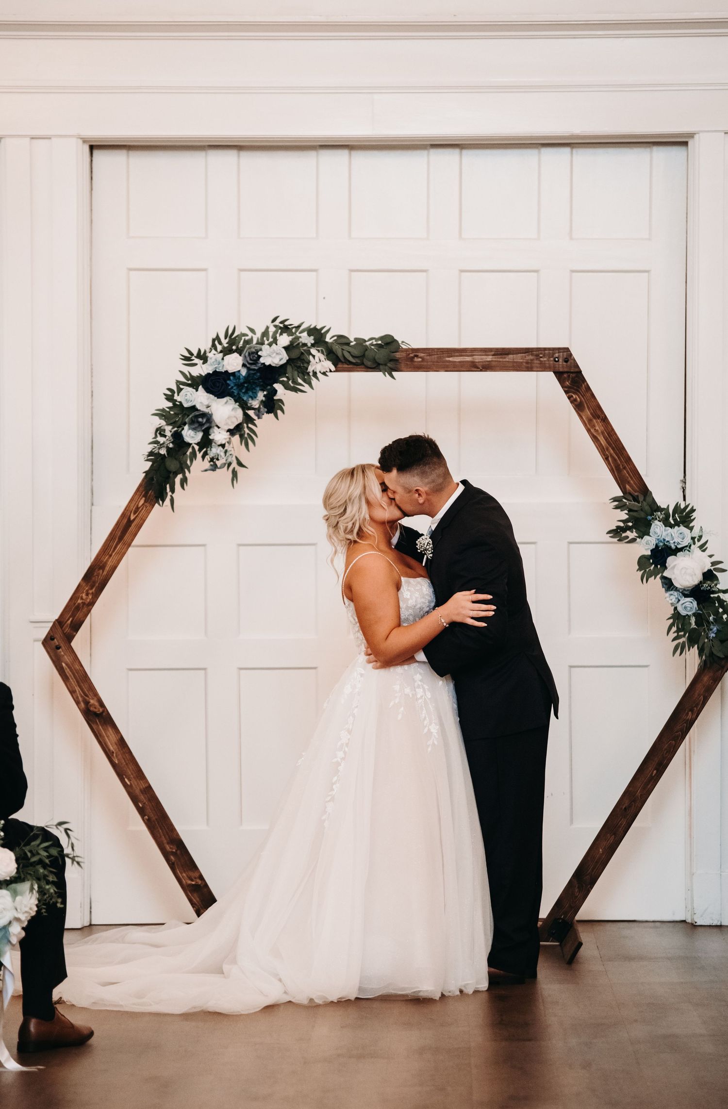 Newlyweds share a romantic moment under a hexagonal wooden arch decorated with white flowers.