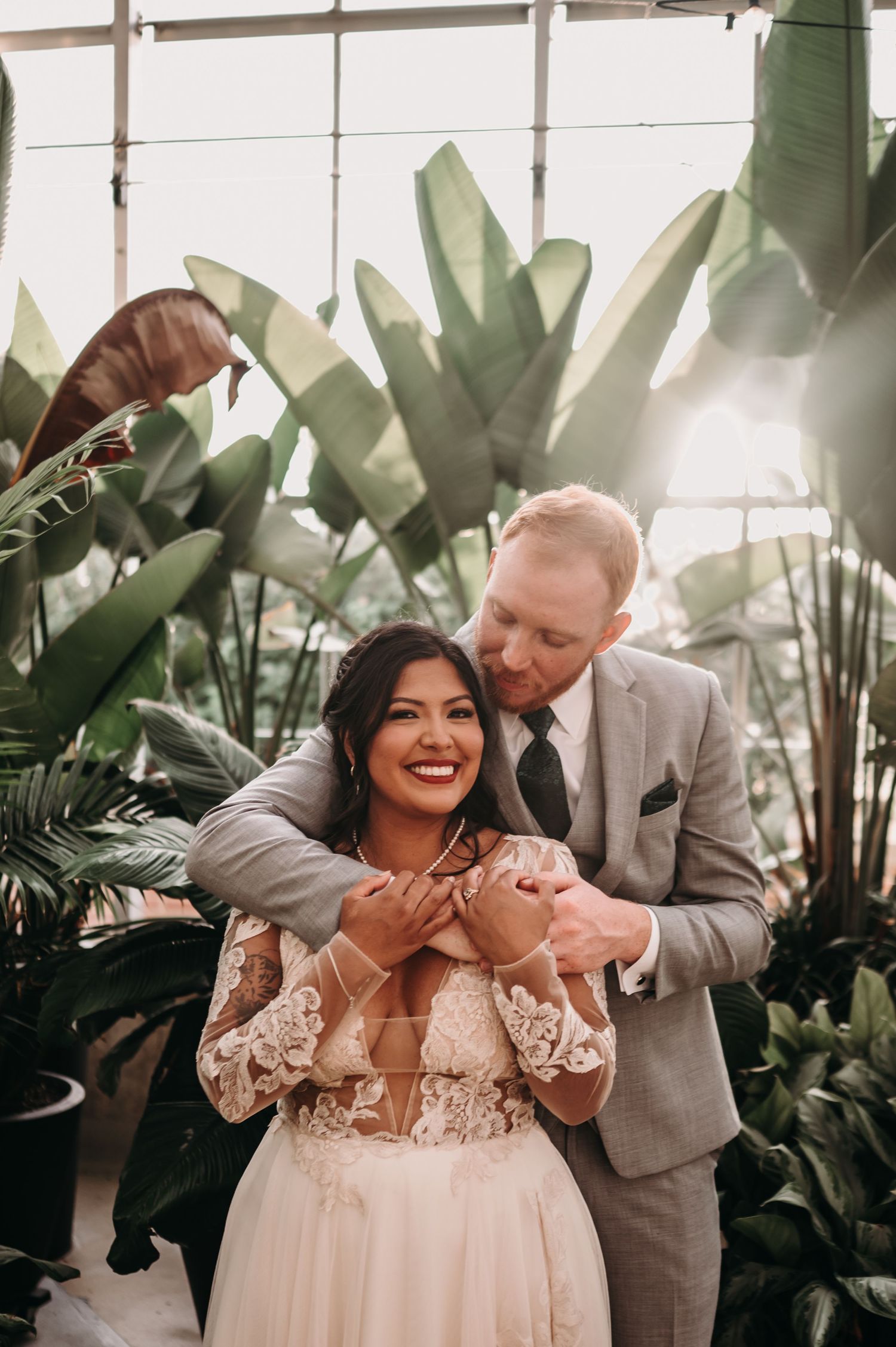 A romantic wedding photo of a couple embracing in a greenhouse surrounded by lush tropical plants.