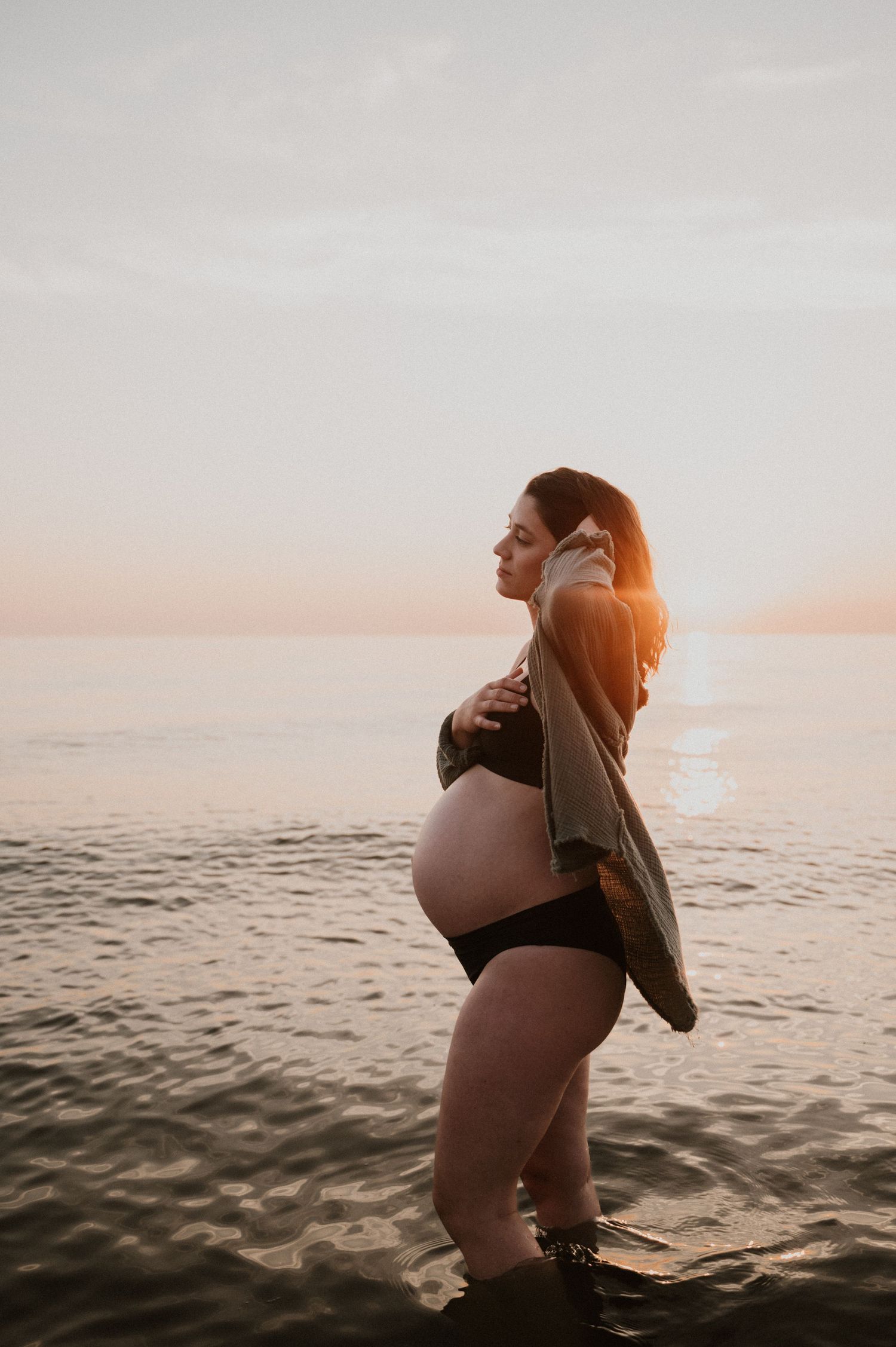 Silhouette of pregnant woman standing in water during sunset wearing a cardigan.