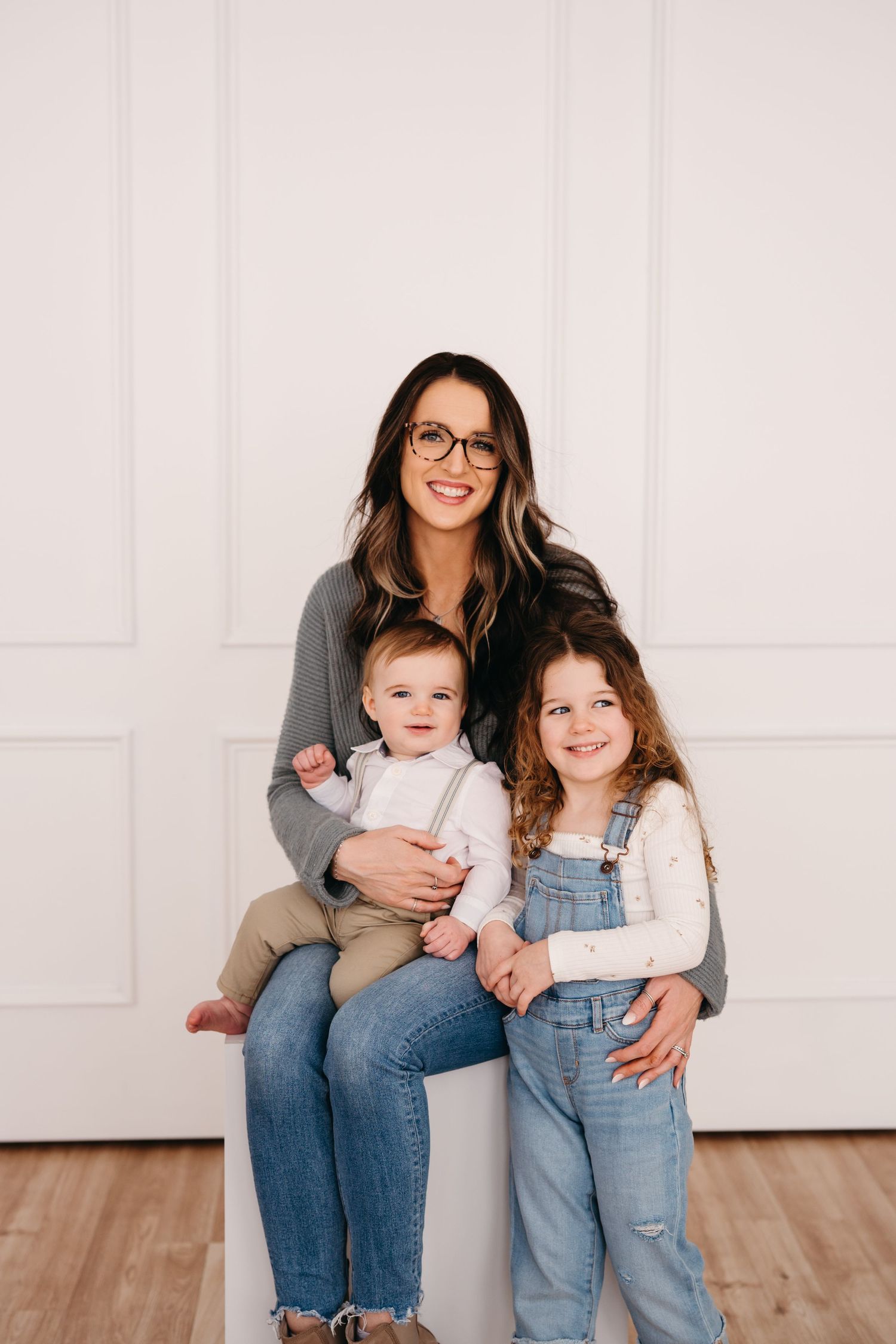 A family portrait featuring a woman in glasses with two children sitting against a white wall.