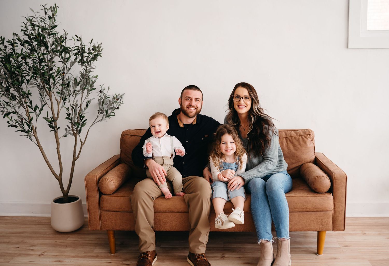 Family of four sitting together on a tan leather couch near a potted olive tree in a bright living room.
