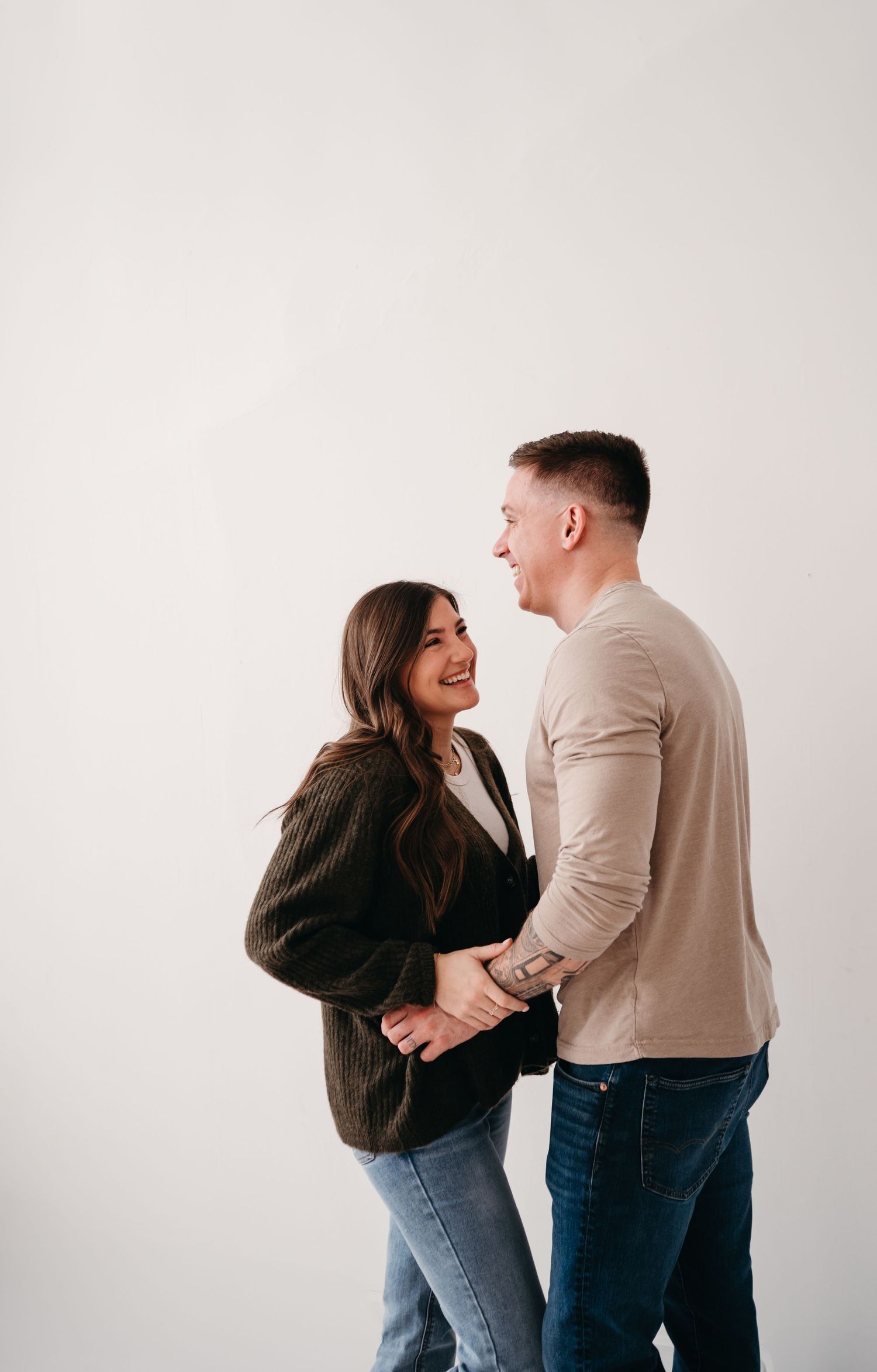 A couple shares a joyful moment together against a white background, dressed casually in sweaters and jeans.