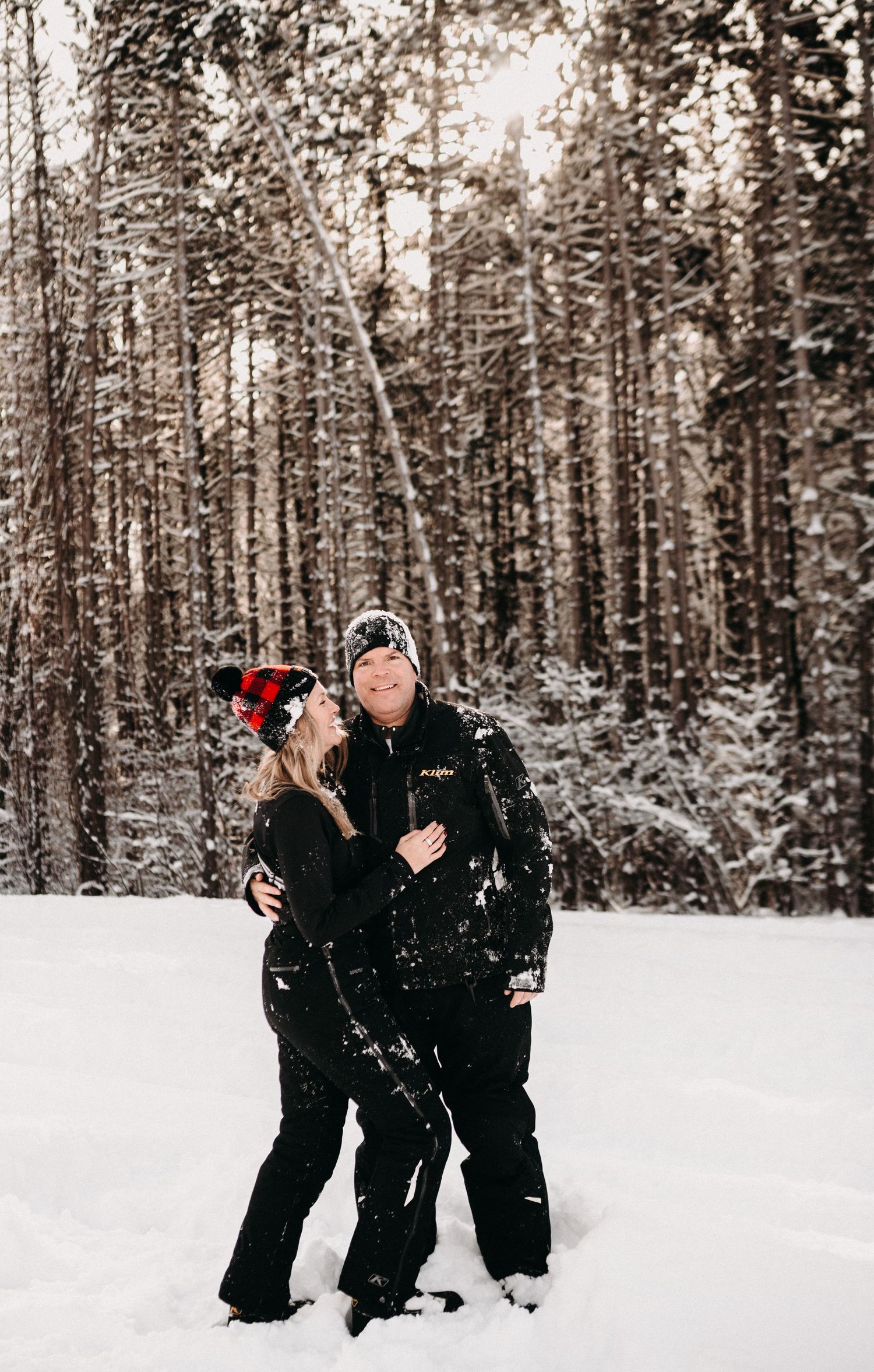 A couple in black winter clothing standing together in a snow-covered forest landscape.