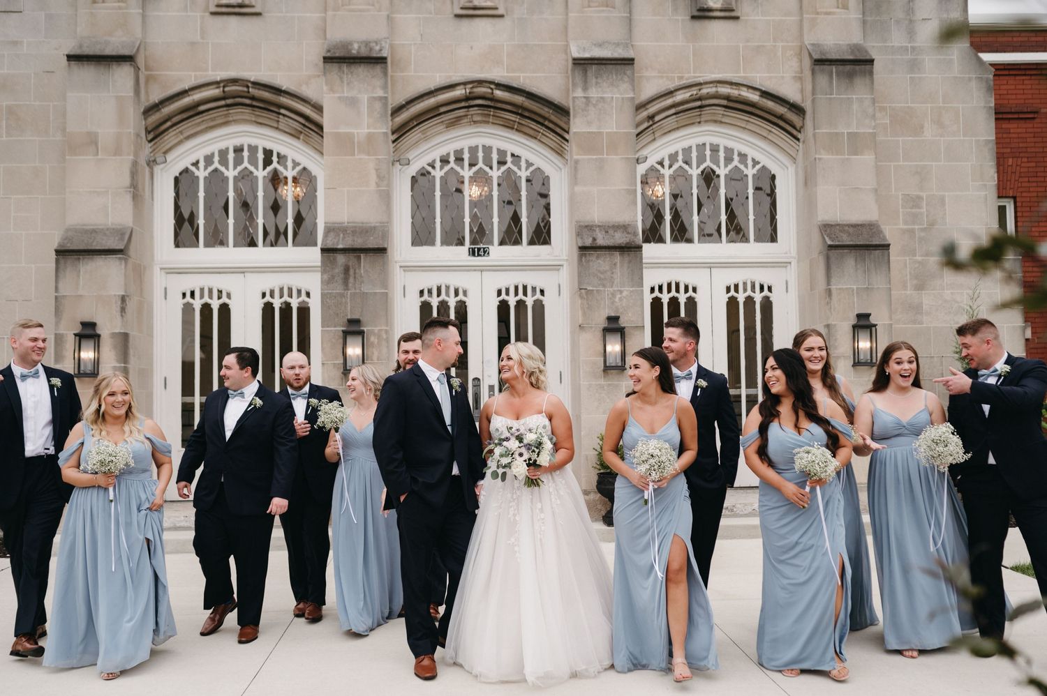 Bridal party laughing together outside elegant stone church with arched windows.