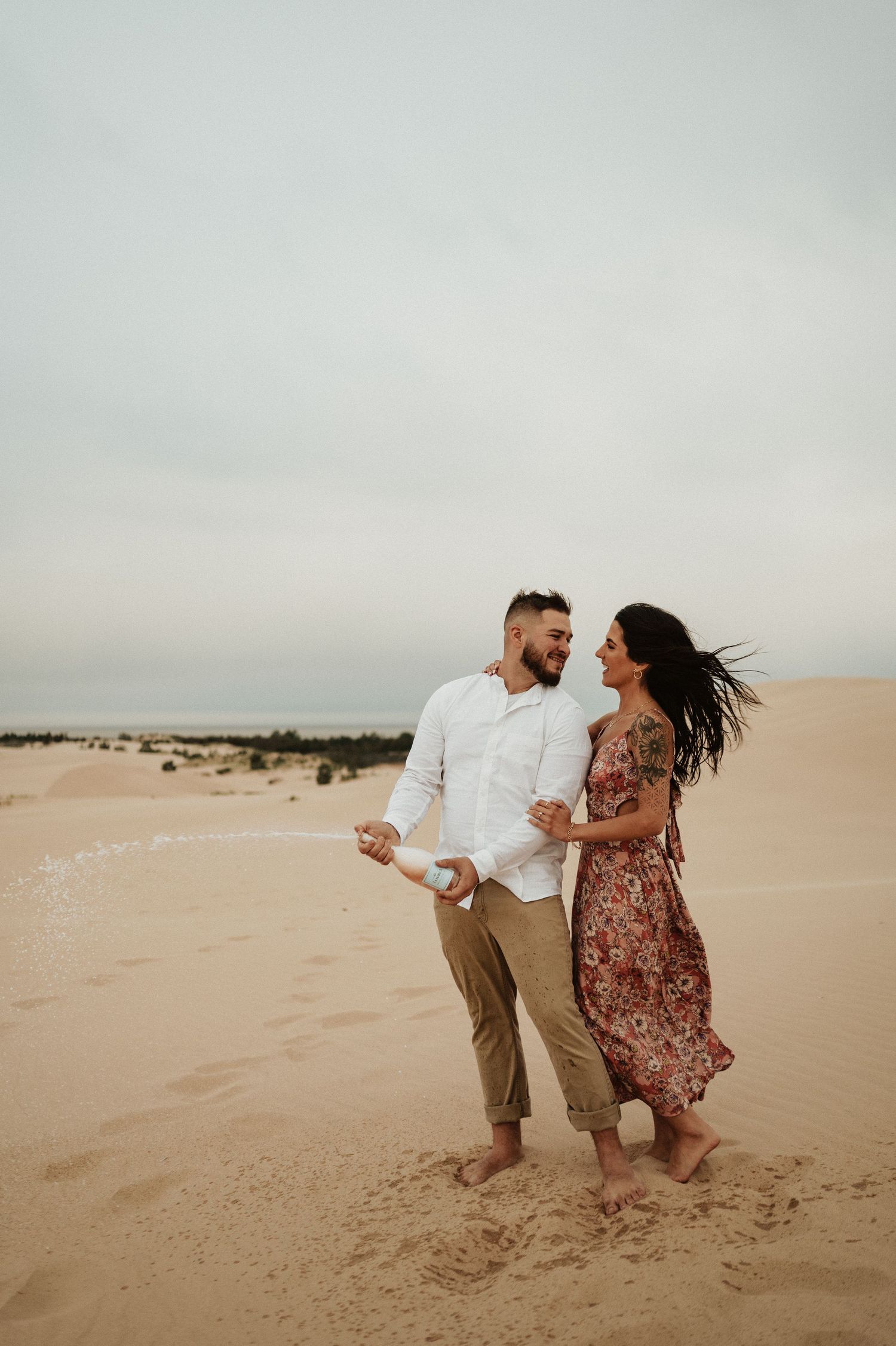 A couple laughs together while walking barefoot across desert sand dunes.