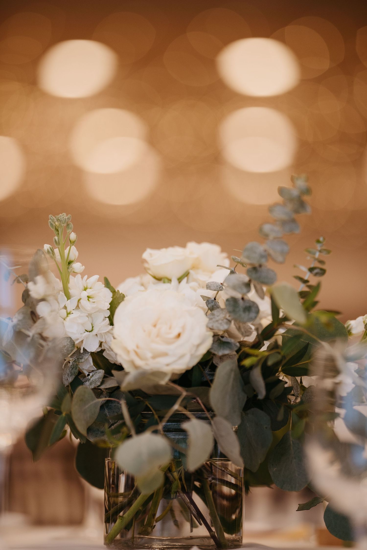 White roses and eucalyptus arrangements in glass vases create an elegant table setting with bokeh lighting.