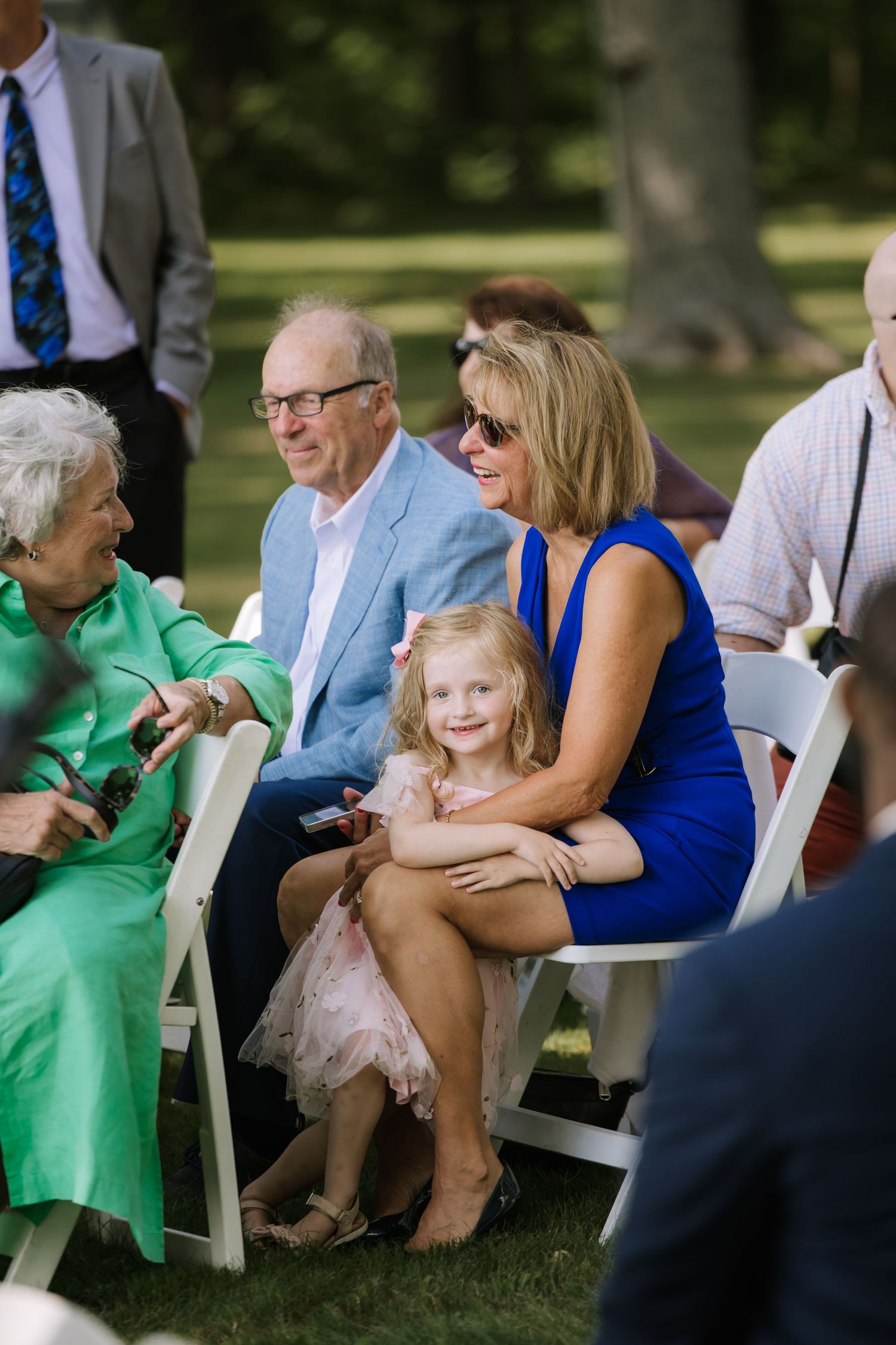 Guests seated in white chairs at an outdoor event with sunlit trees in the background.