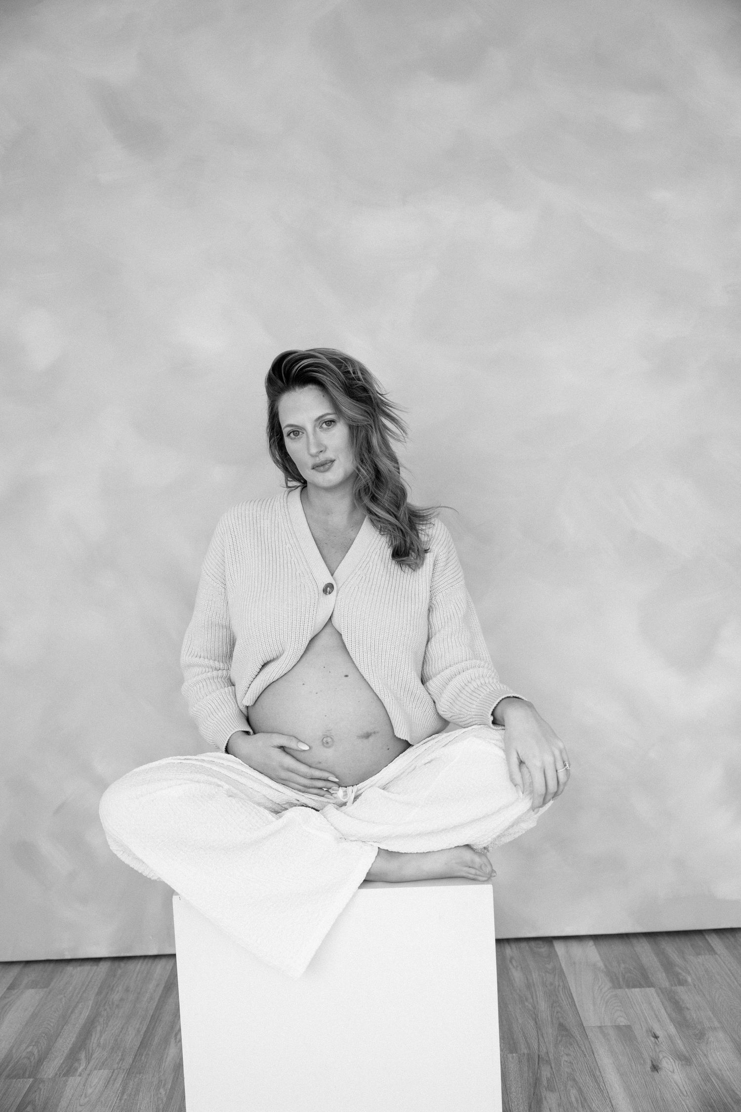 Black and white portrait of pregnant woman in cardigan sitting cross-legged on white cube, cradling her bare bump.