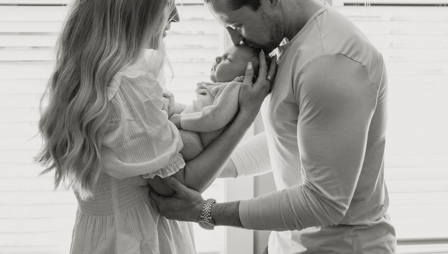 Black and white photo of a couple tenderly holding their newborn baby together in a bright, softly lit room.