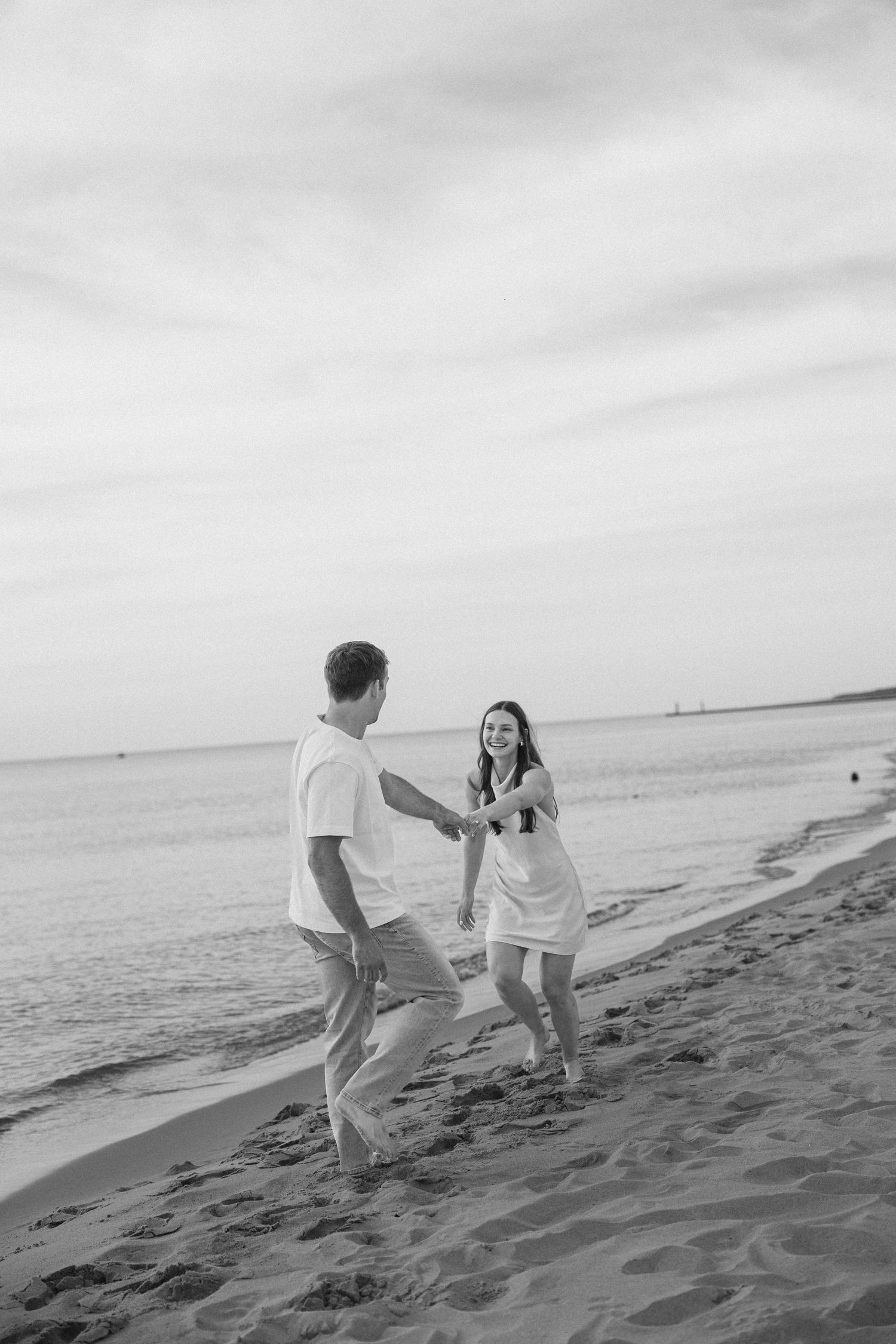 Black and white photo of joyful couple dancing and holding hands on a serene beach at sunset.