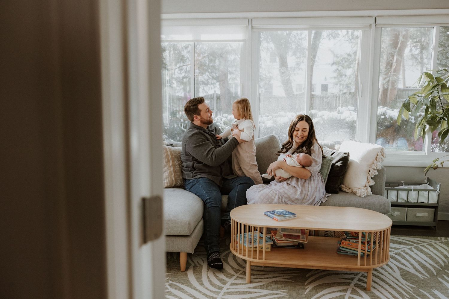 Cozy in-home family photo session with parents and newborn baby by bright sunlit windows.