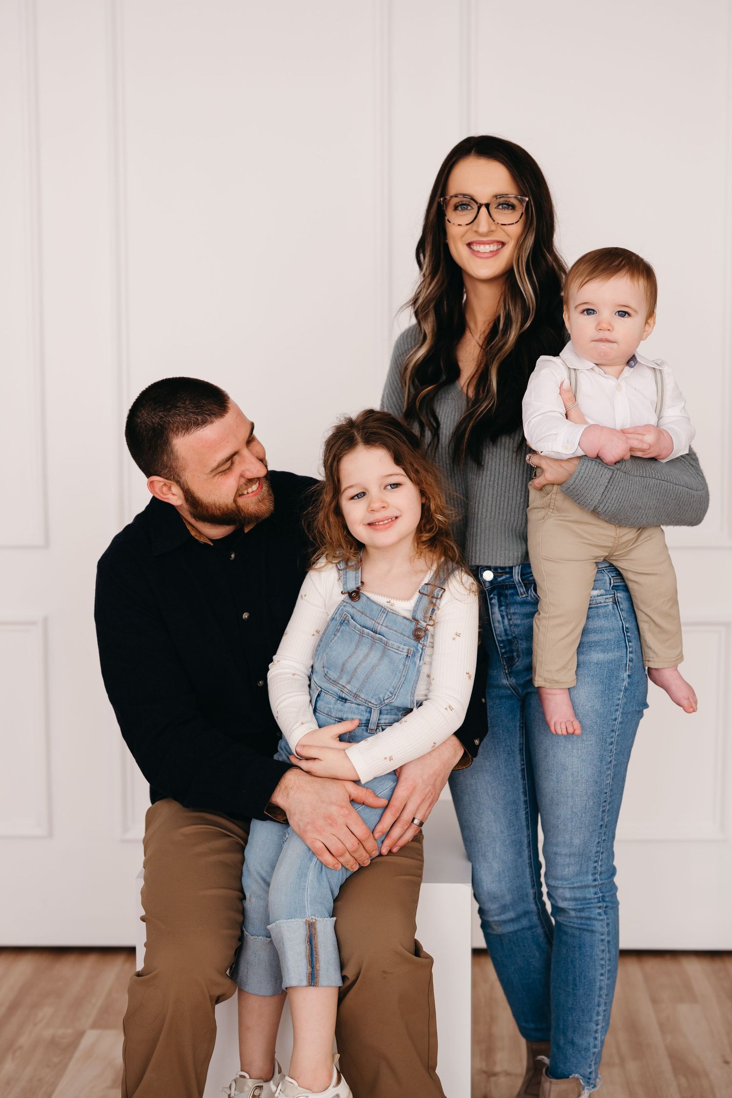 A family of four poses together against a white wall on hardwood flooring.