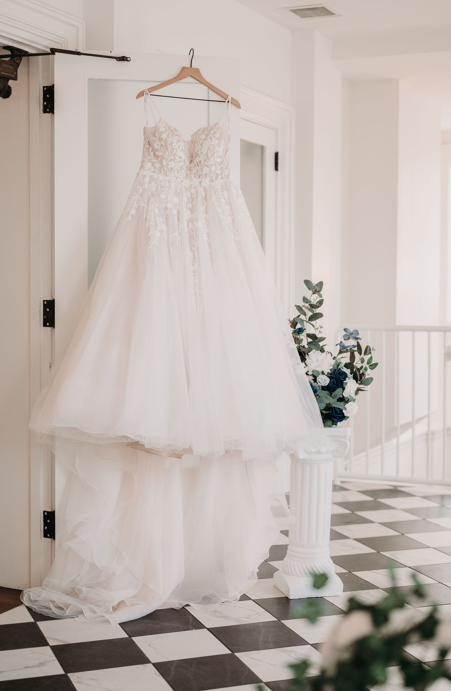 A delicate white wedding dress with lace details hangs on display against a white wall.
