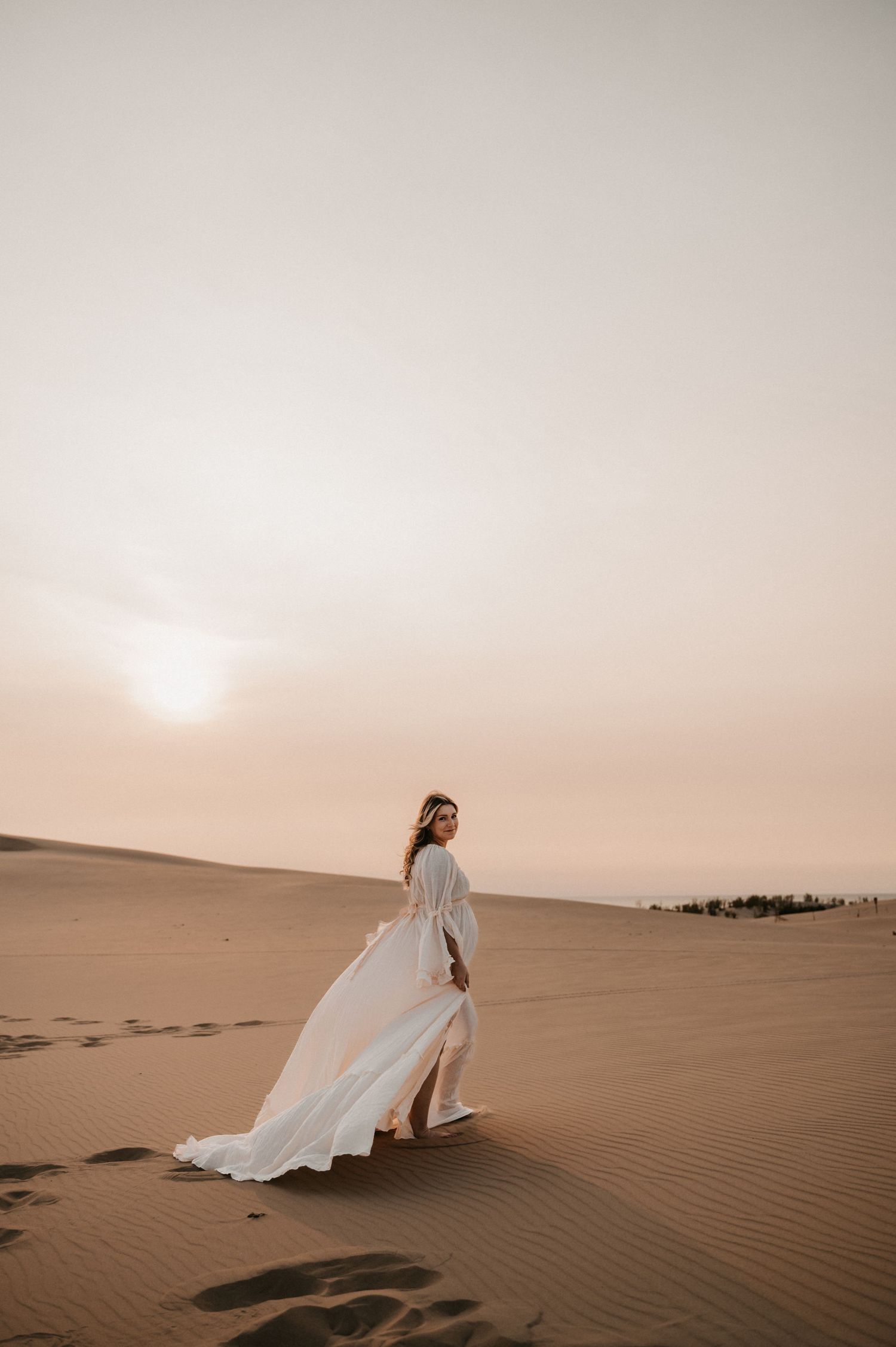 A flowing white dress catches the desert wind as a woman stands on a sand dune at sunset.