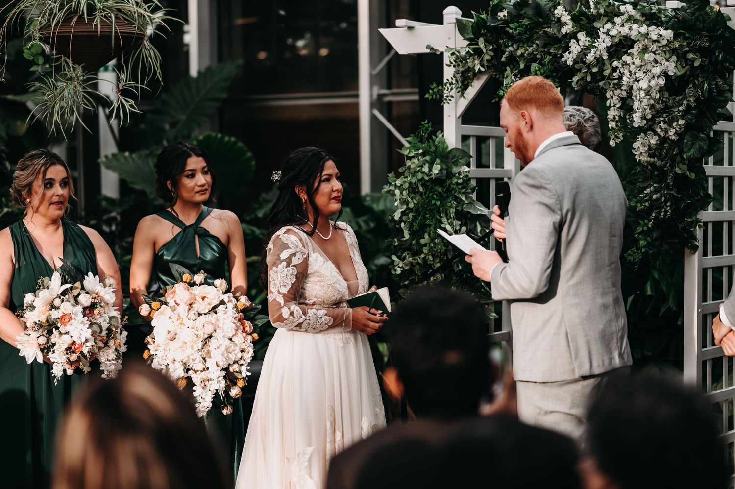 Exchange of vows during greenhouse wedding ceremony surrounded by tropical plants and string lights.