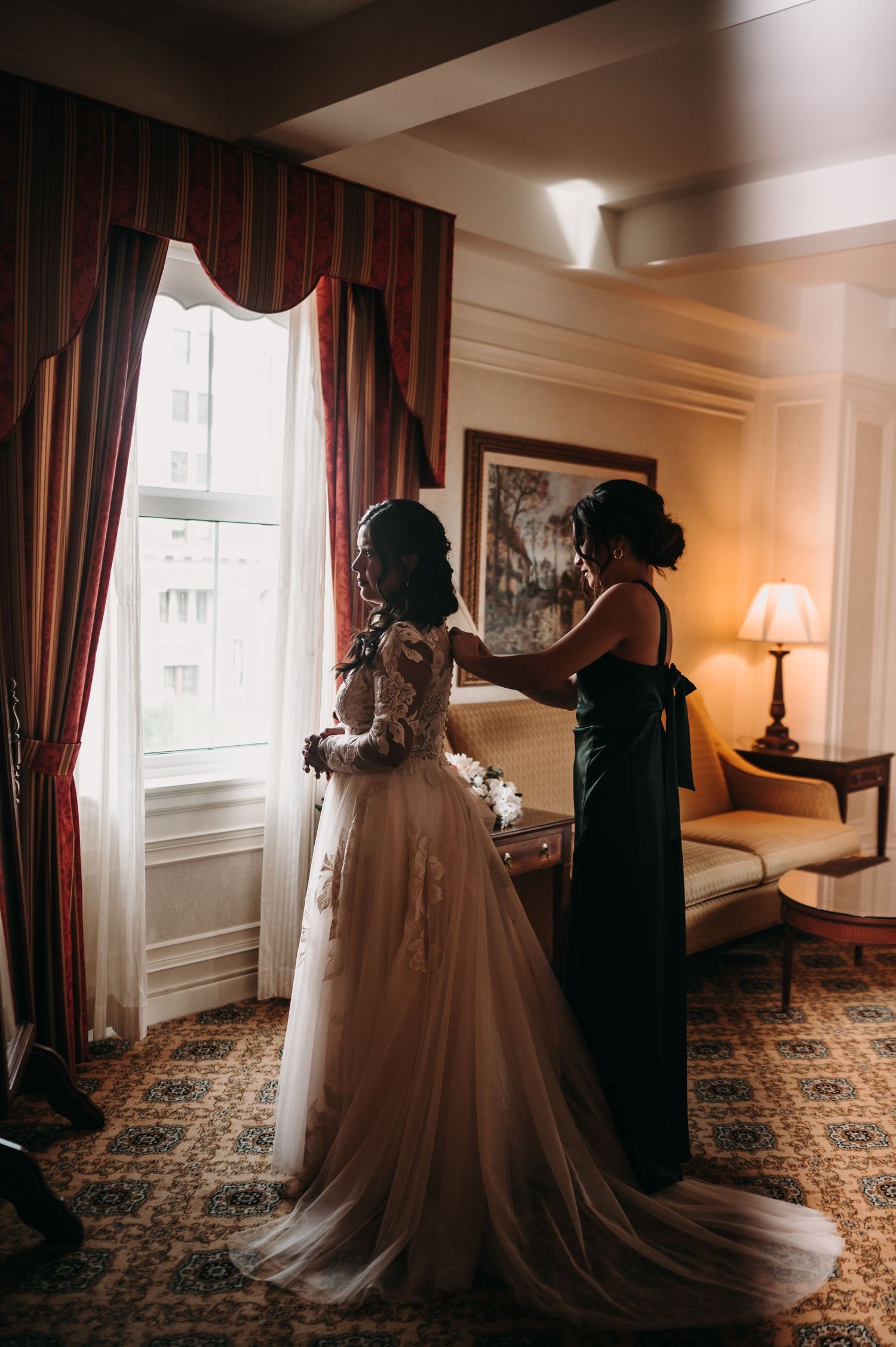 A wedding dress fitting session in an elegant room with dark red curtains and patterned carpet.