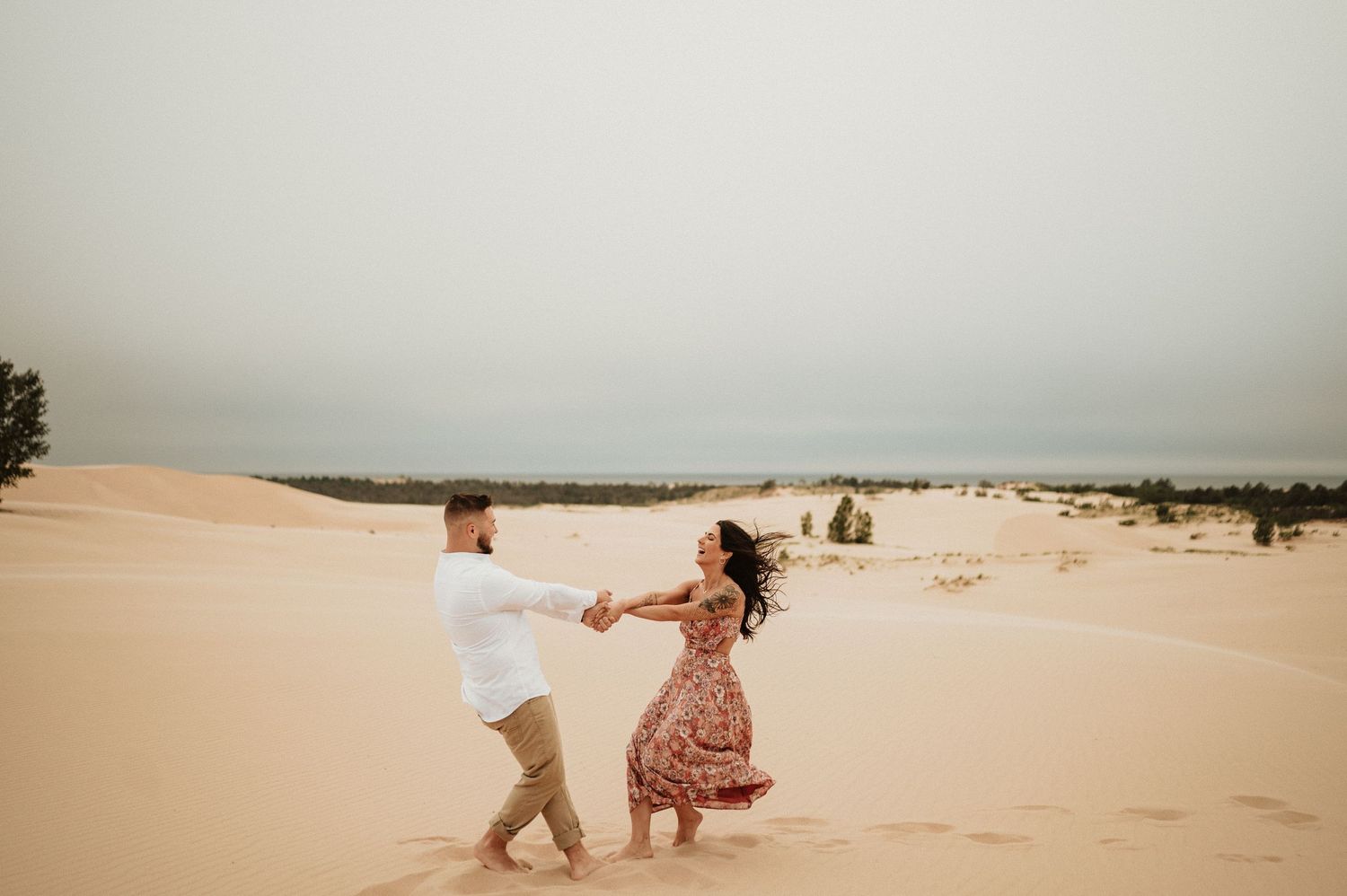 A couple dances playfully together on sandy dunes under an overcast sky.