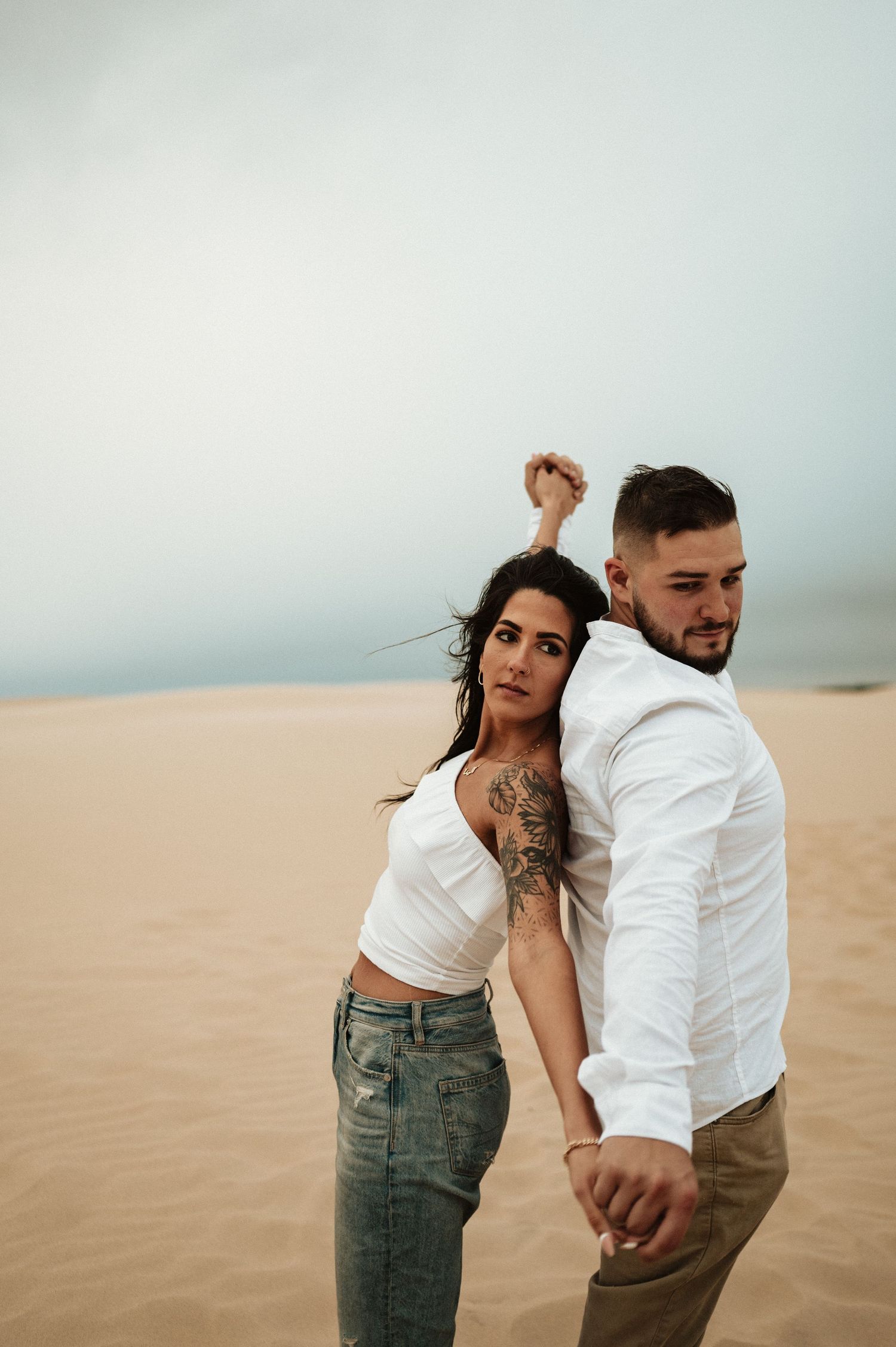 A couple in white and denim poses back-to-back on sandy dunes during a photoshoot.
