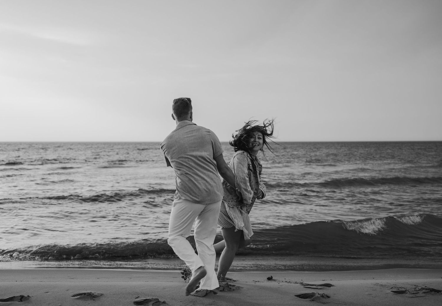 Couple walking along a serene beach shoreline at sunset in black and white.