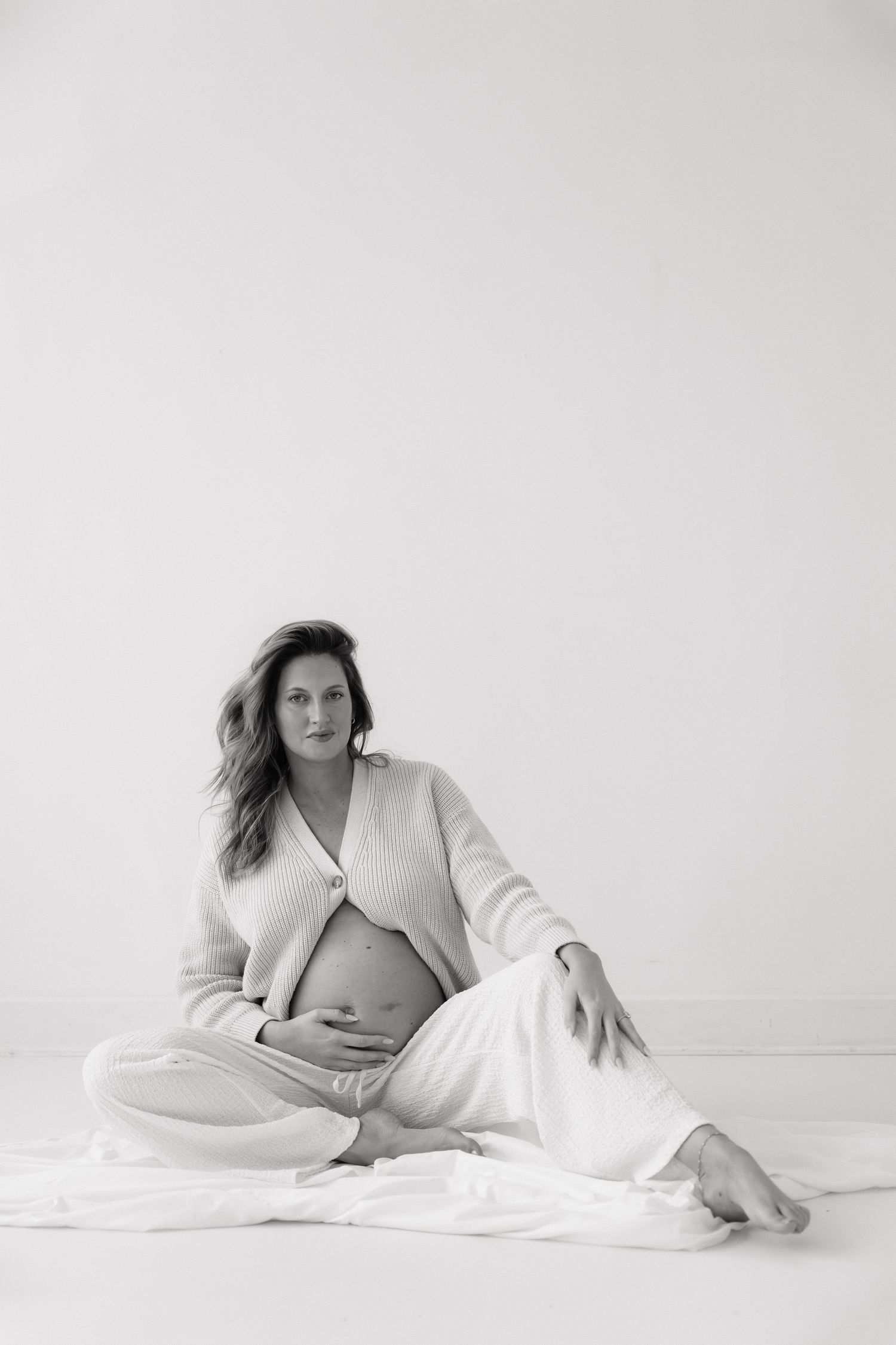 Black and white photo of a pregnant woman sitting cross-legged on the floor, cradling her bare baby bump in a knit cardigan.
