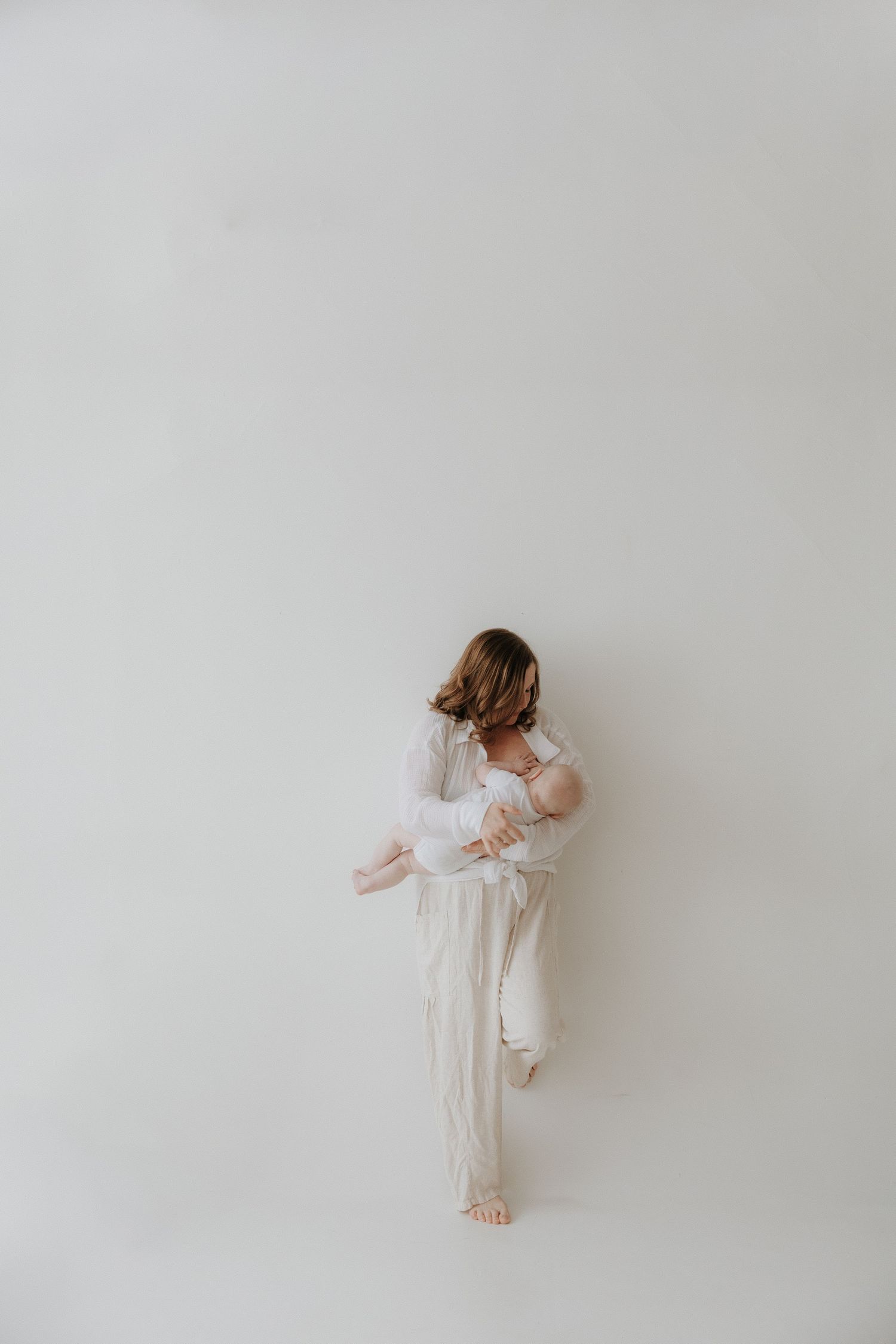 A mother in white linen holds her newborn baby while standing against a light gray studio background.